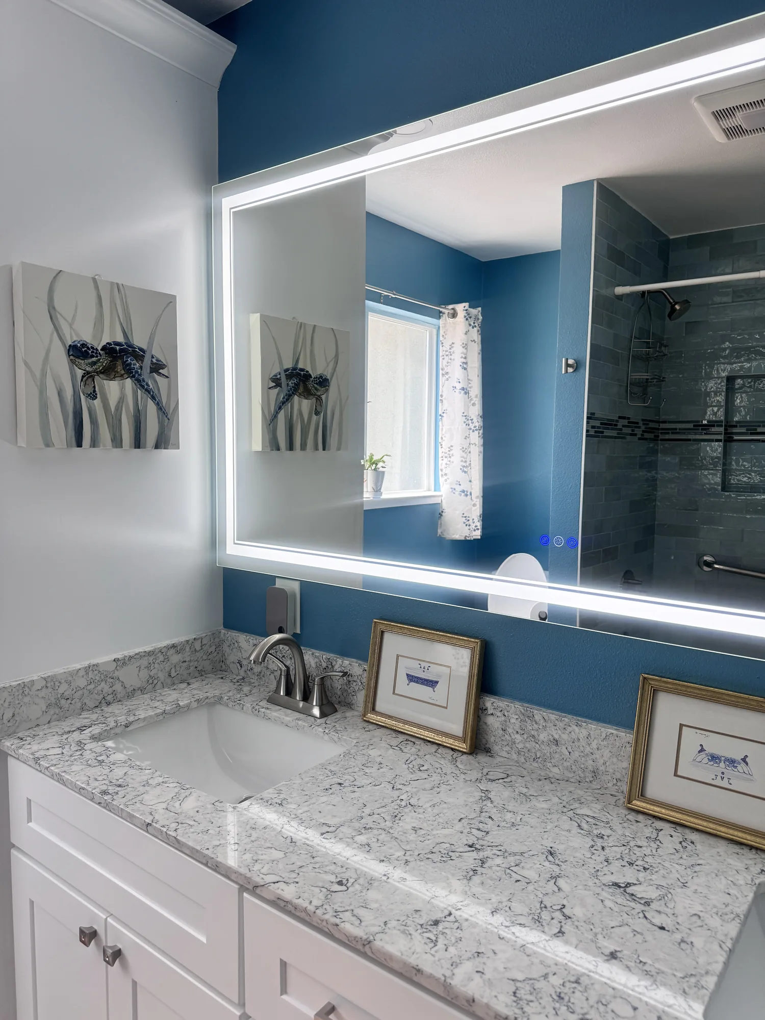 Backlit mirror above the stone-look vanity top with the tiled shower reflected in the glass.
