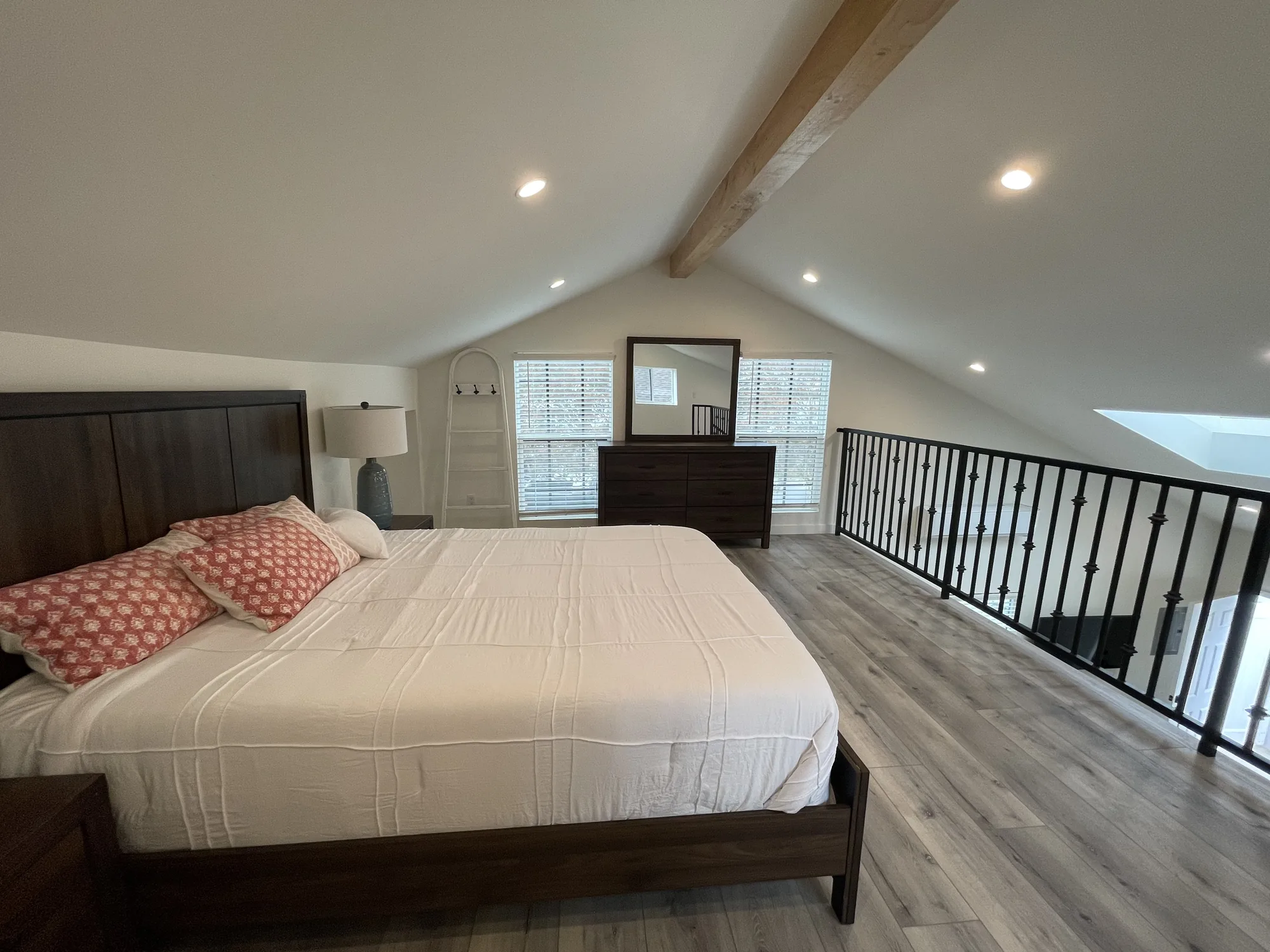 Wide loft bedroom with vaulted ceiling, wood beam, iron railing along the open edge and a skylight.