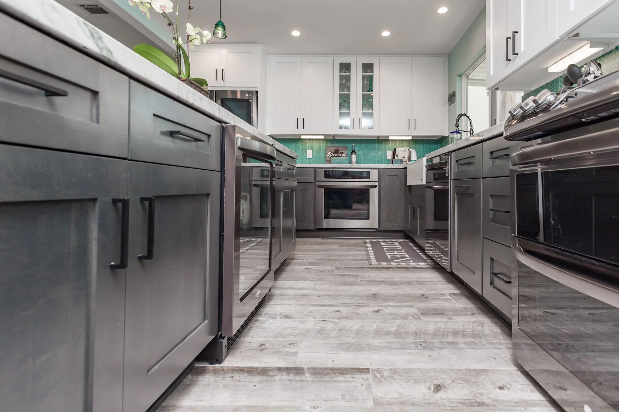 Floor-level view down the kitchen showing gray cabinets, plank flooring, and stainless appliances.