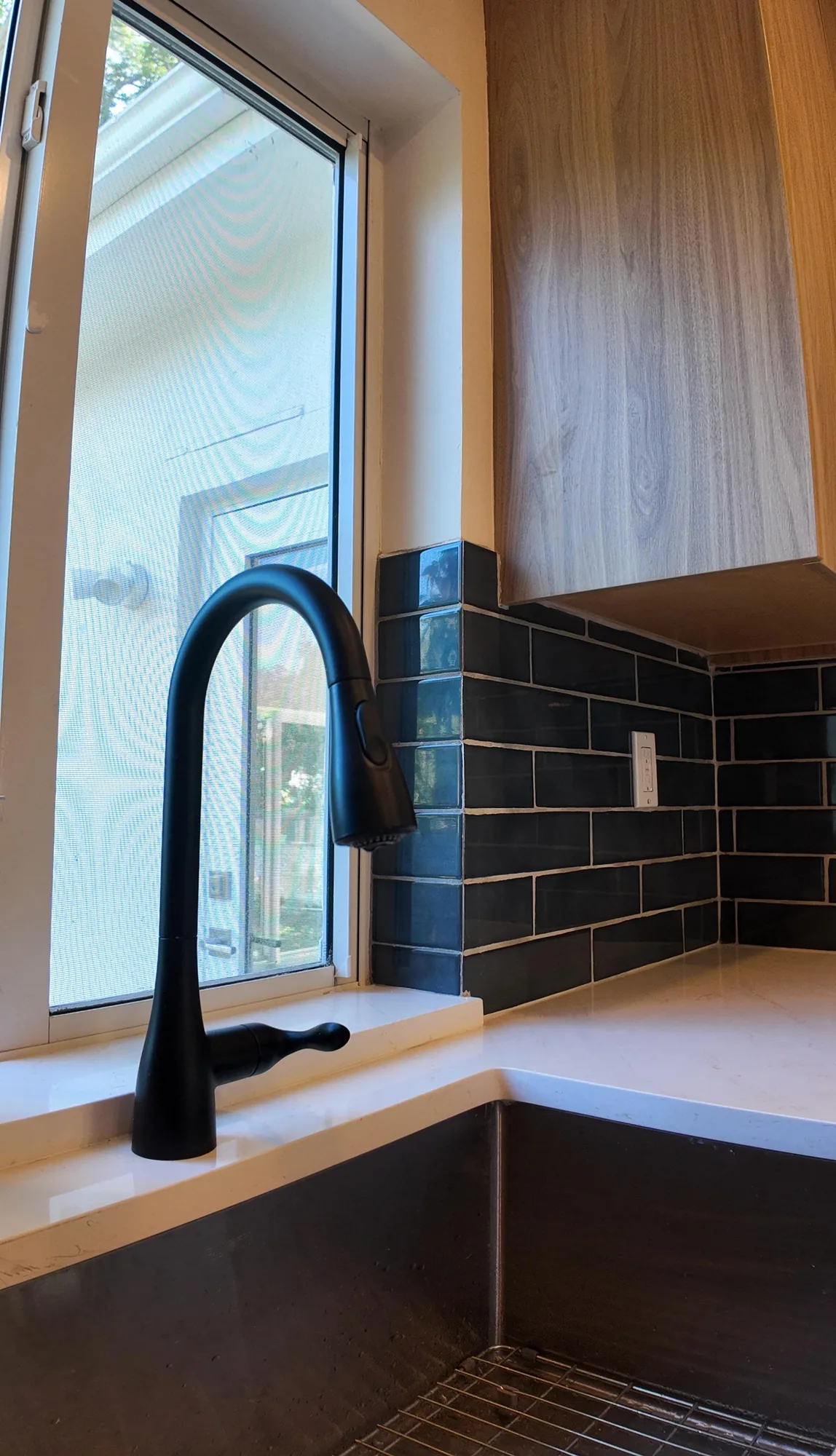 Close view of a matte black pull-down kitchen faucet beside a window and dark glossy subway tile.