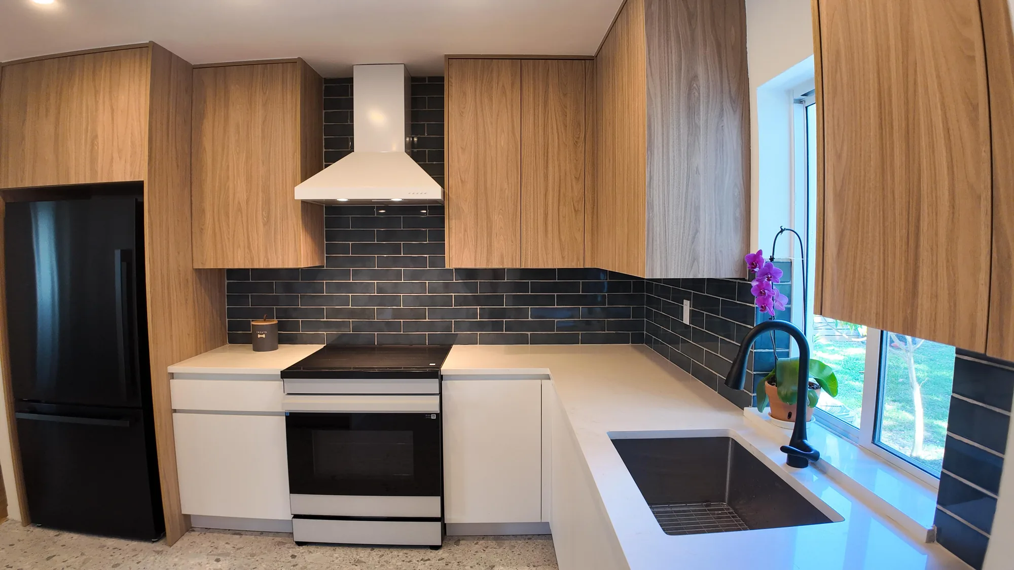 Wide corner view of a modern kitchen with wood upper cabinets, white lowers and dark tile backsplash.