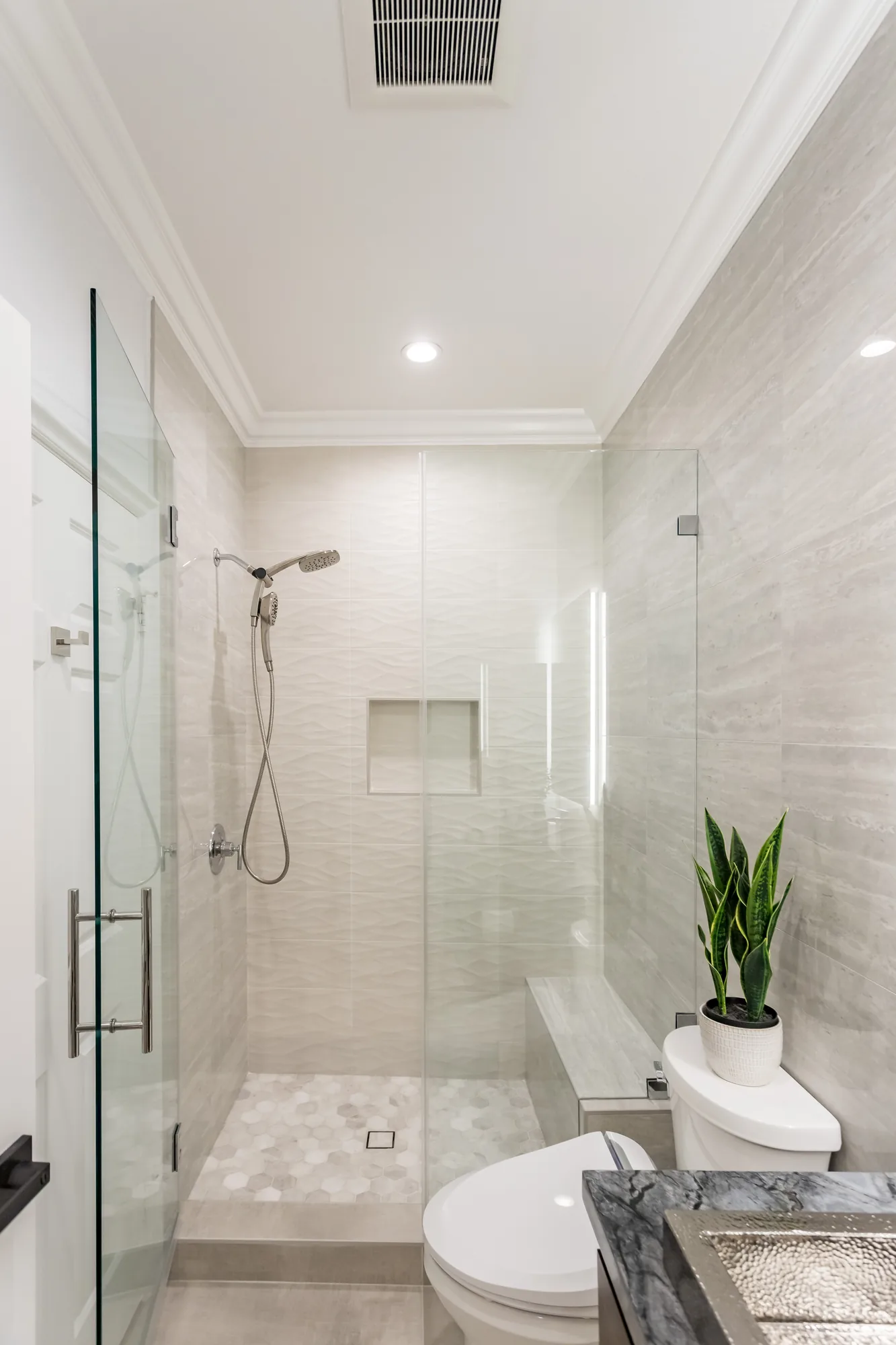 Vertical view of a narrow bathroom with a frameless glass shower, tile bench and hexagon mosaic floor.