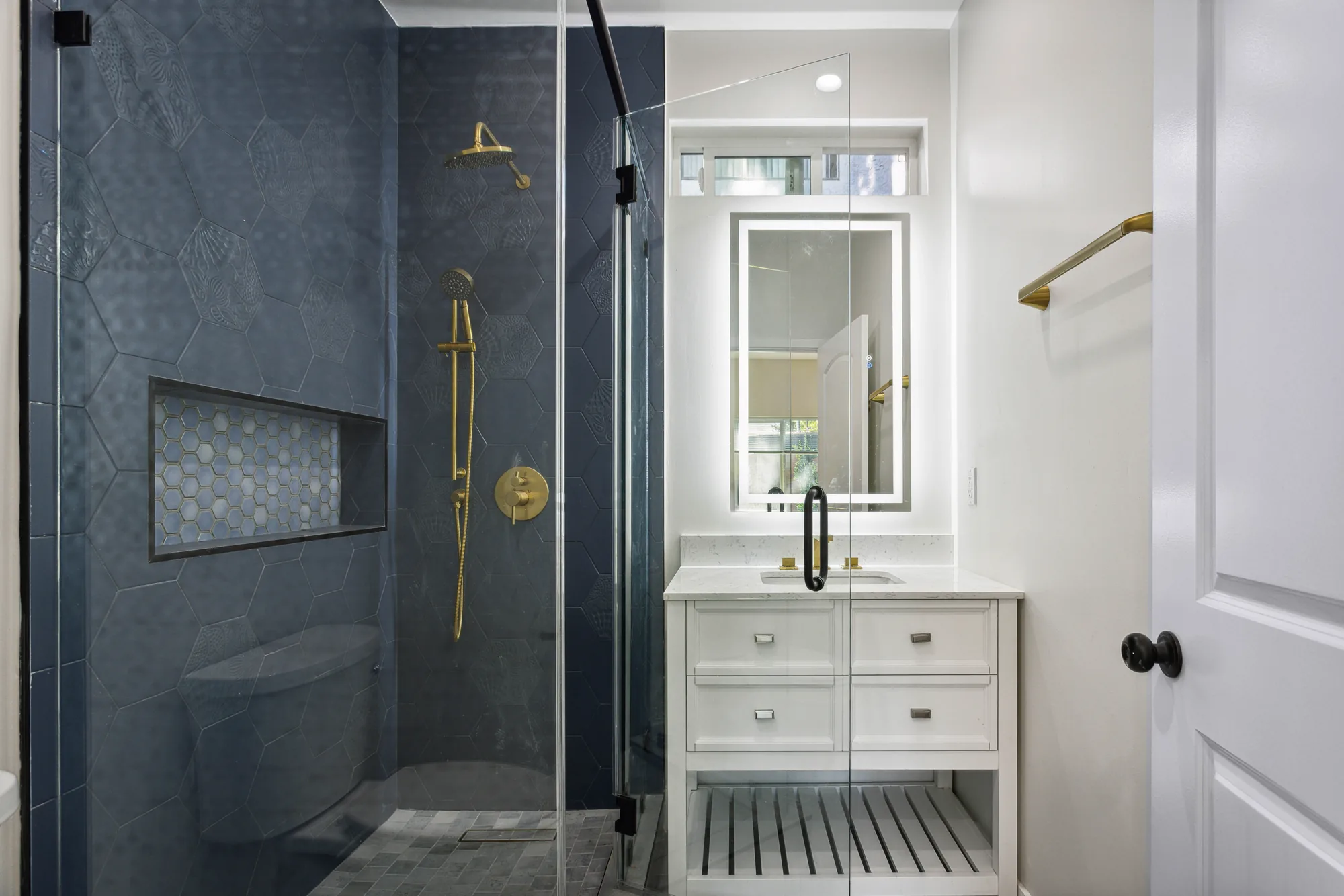 Bathroom with a navy hexagon tile shower behind glass and a white vanity with a lighted mirror.