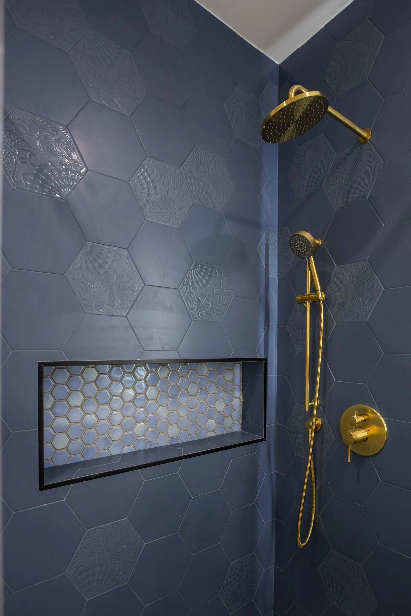 Navy hexagon shower tile with raised pattern accents, brass rain head, handheld and niche.