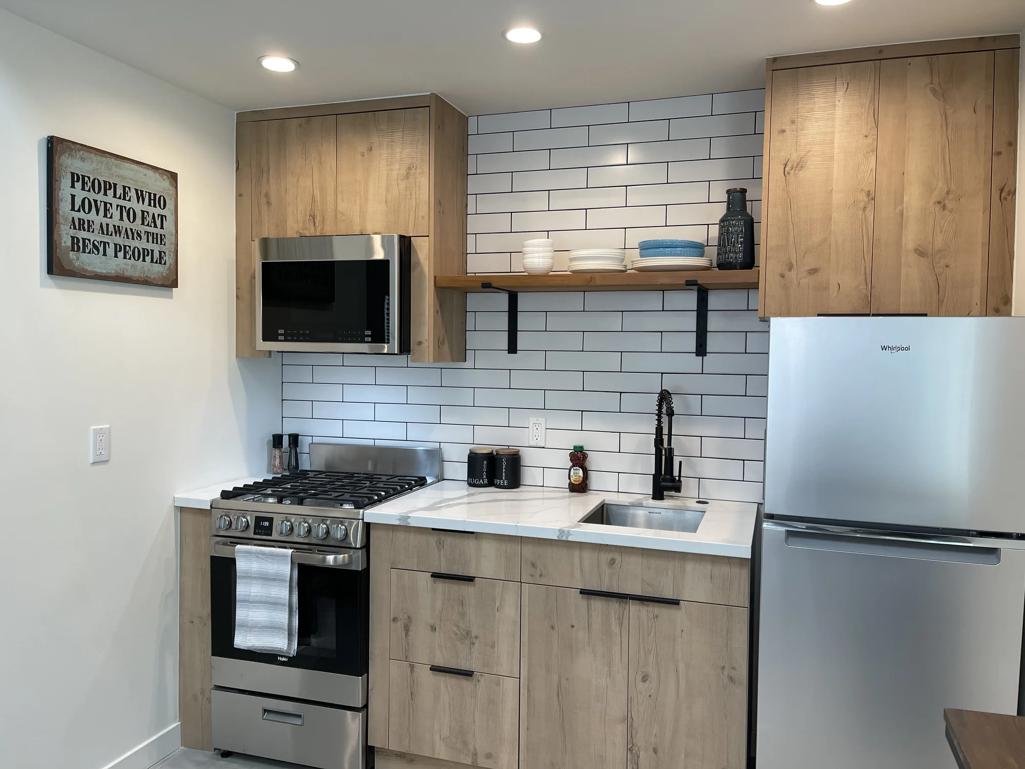Compact kitchen with oak-look slab cabinets, white subway tile and a floating wood shelf.