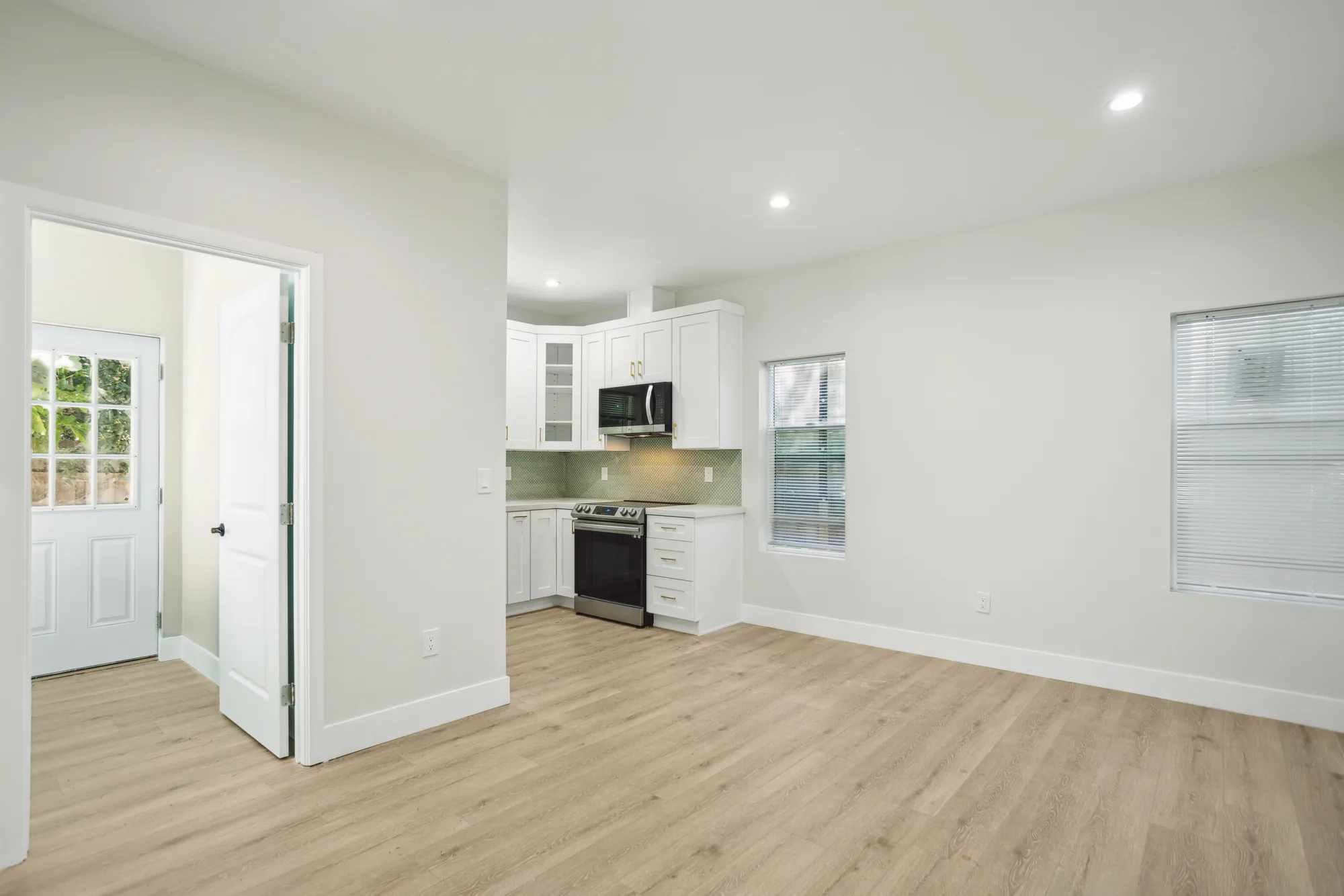 Open living area with light wood-look plank floors looking toward a compact white kitchen.