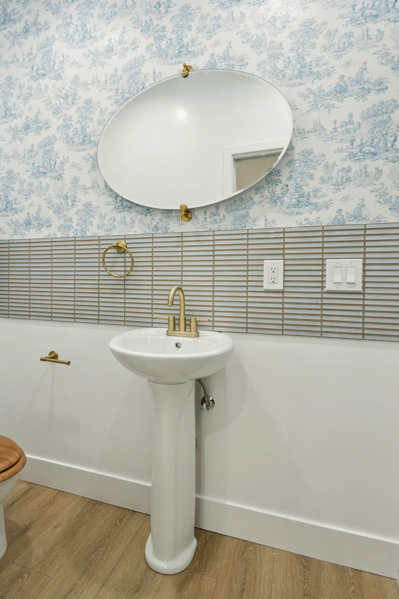 Pedestal sink with a brass faucet below an oval mirror against blue patterned wallpaper.