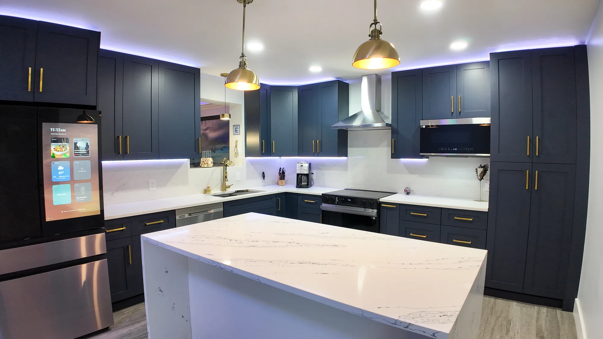 White quartz island with grey veining anchors a navy blue kitchen lit by brass pendant lights.