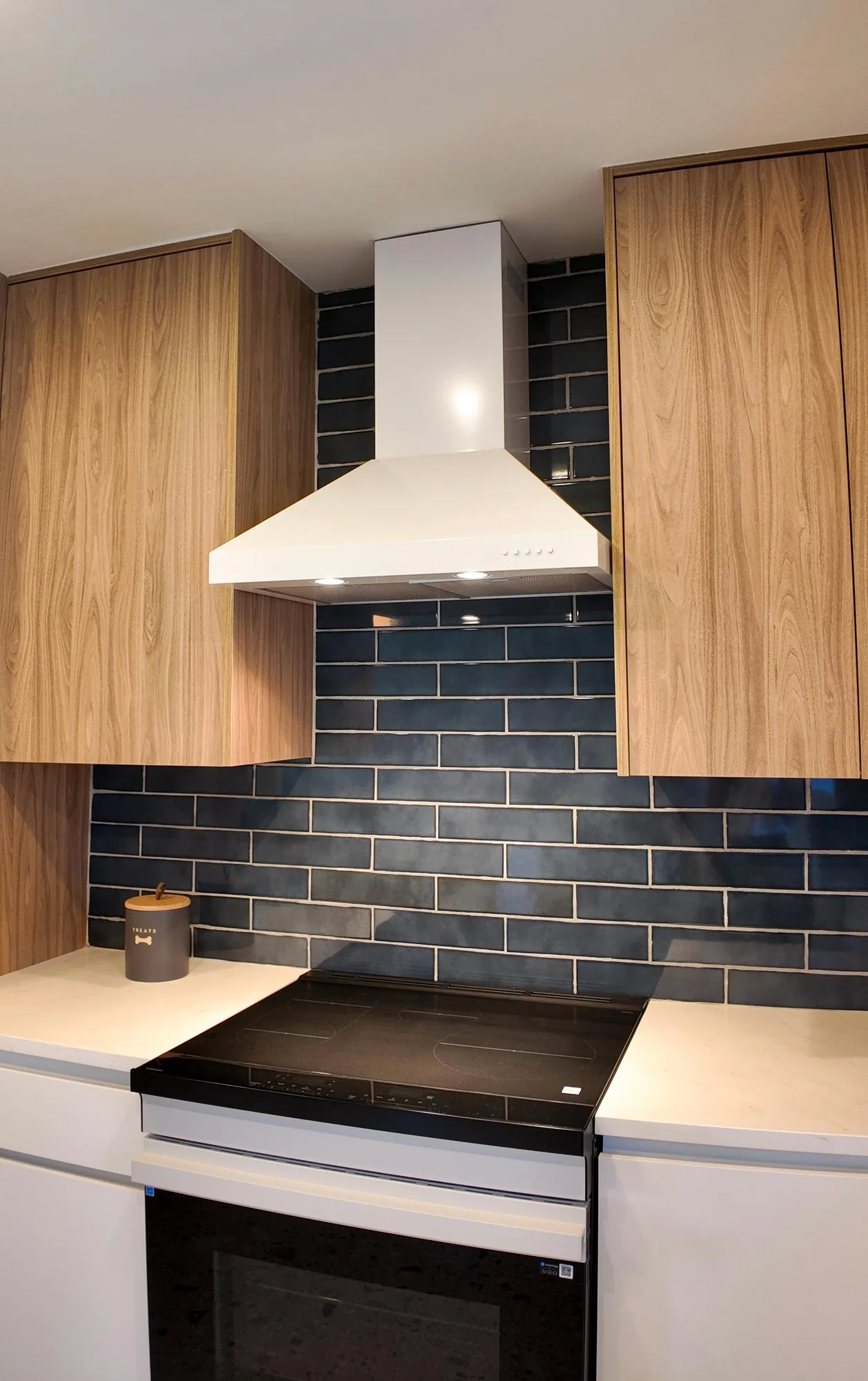 White chimney hood above a smooth-top range, framed by dark subway tile and wood veneer cabinets.