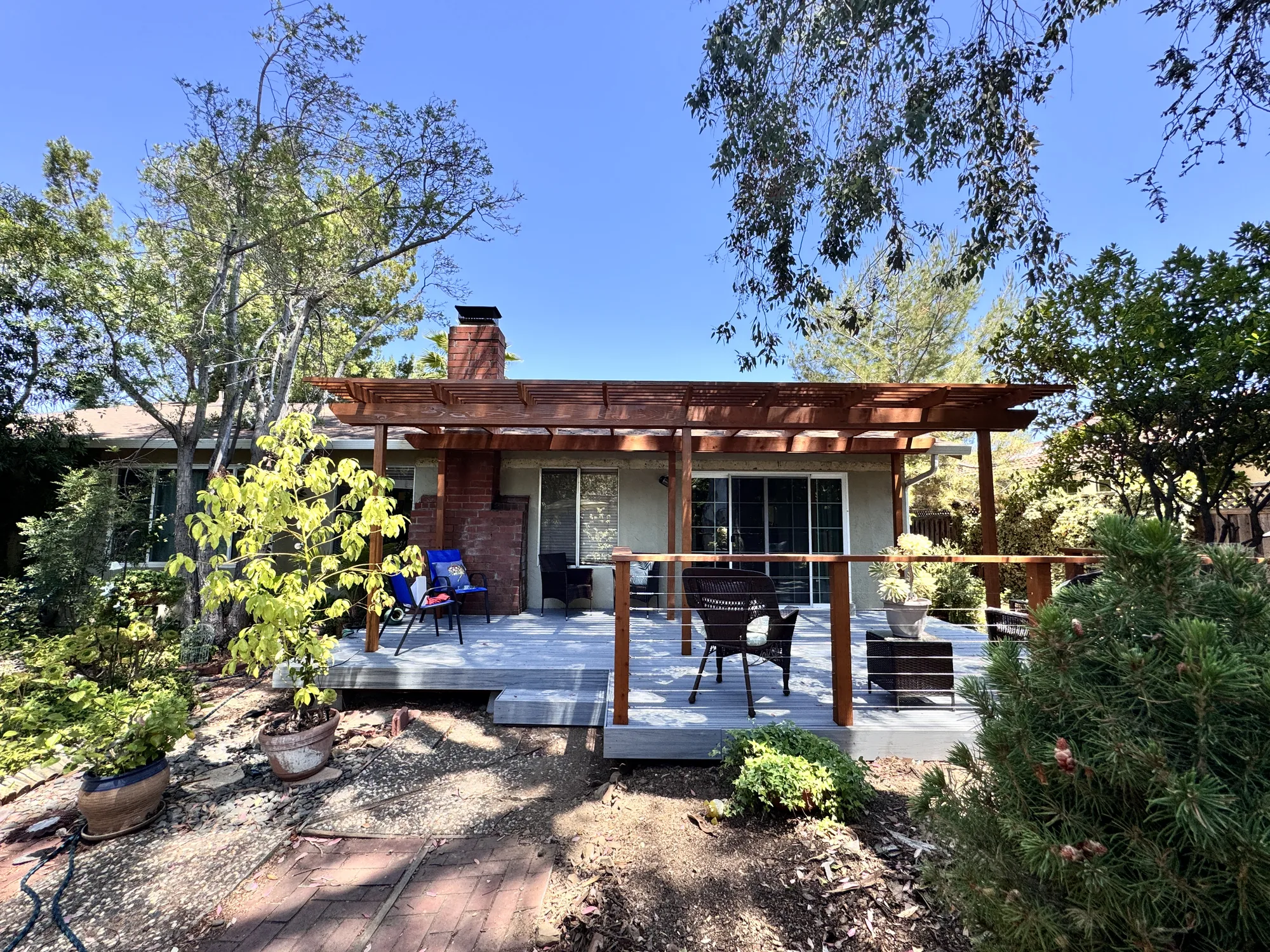 Rear elevation of a home with a new gray deck, wood pergola and cable railing framed by mature trees.