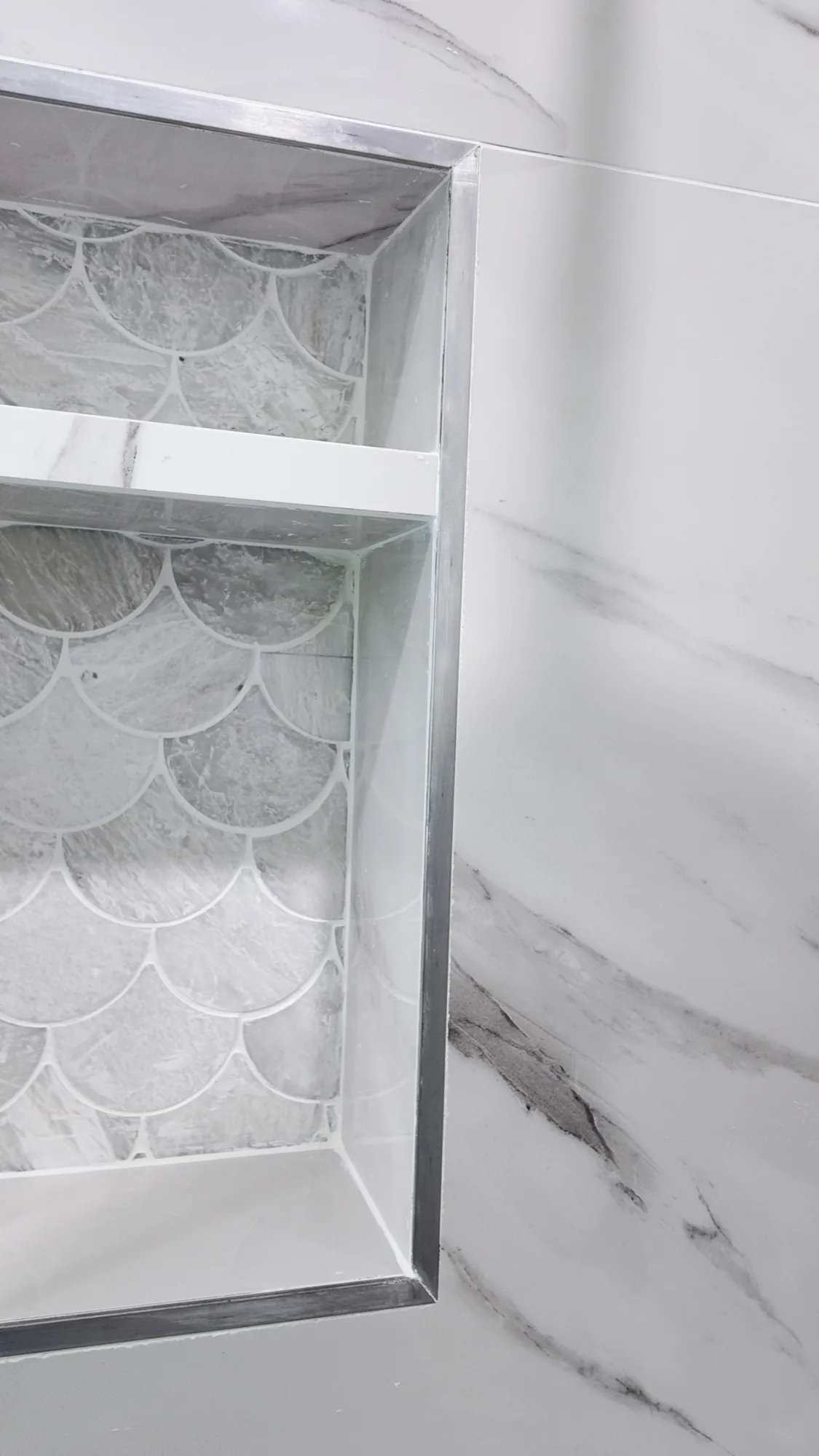 Close view of a recessed shower niche with a shelf, metal edge trim and scalloped stone mosaic back.