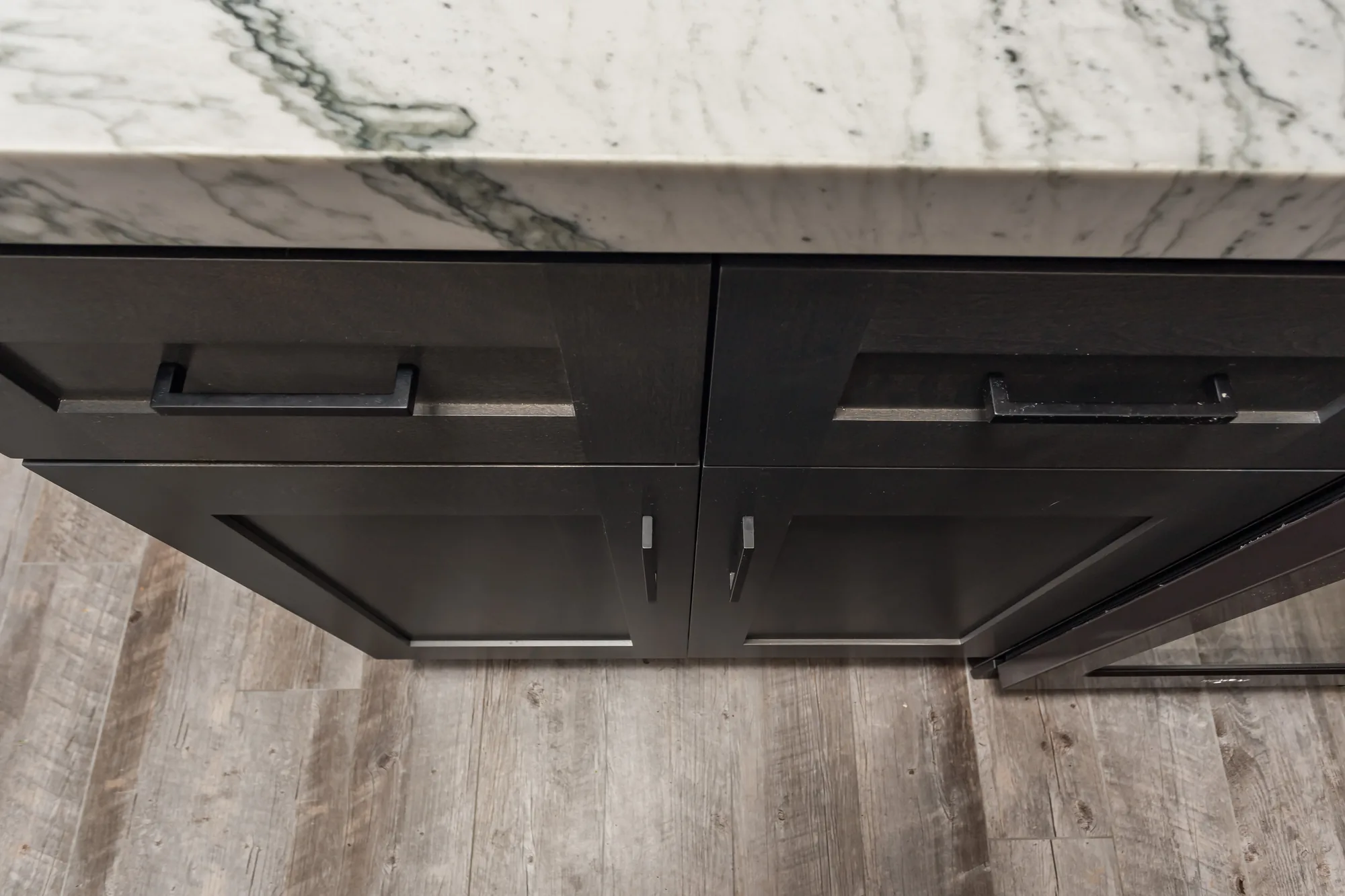Close-up of a gray shaker drawer and door with matte black bar pulls beside plank flooring.