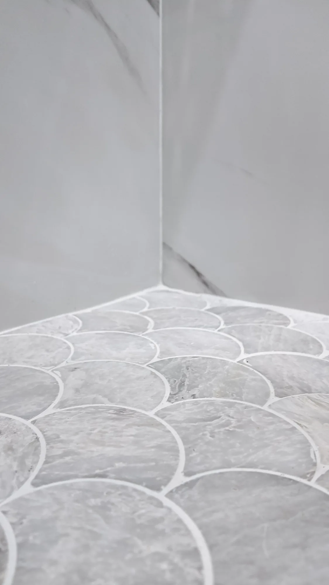 Corner of a tiled shower where scalloped grey stone mosaic flooring meets marble-look wall panels.