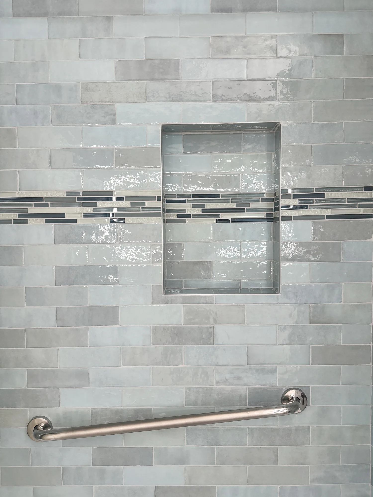 Straight-on view of a recessed tiled niche with a mosaic stripe and grab bar below it.