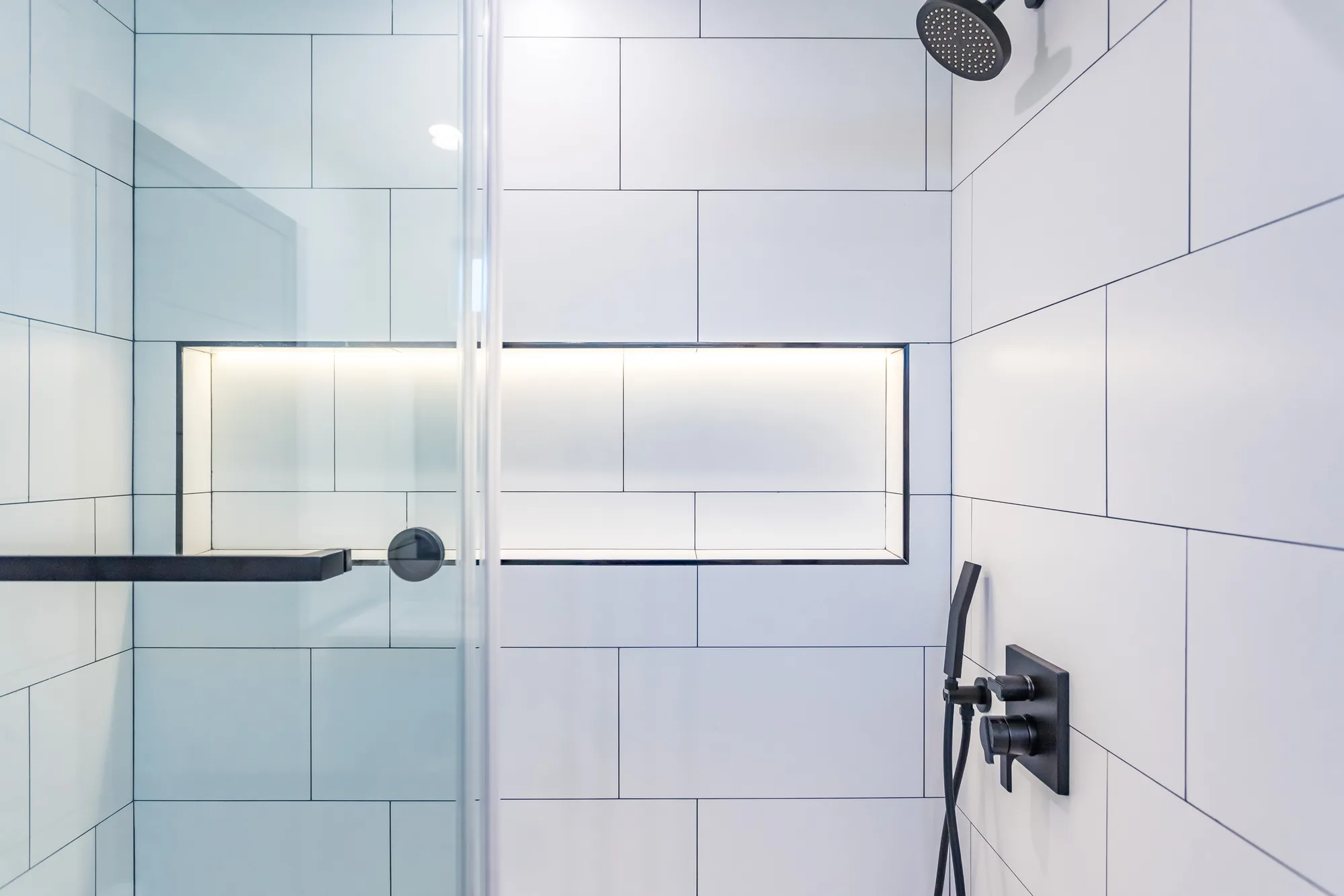 Shower wall with a lit niche, black shelf and matte black valve trim on white large-format tile.