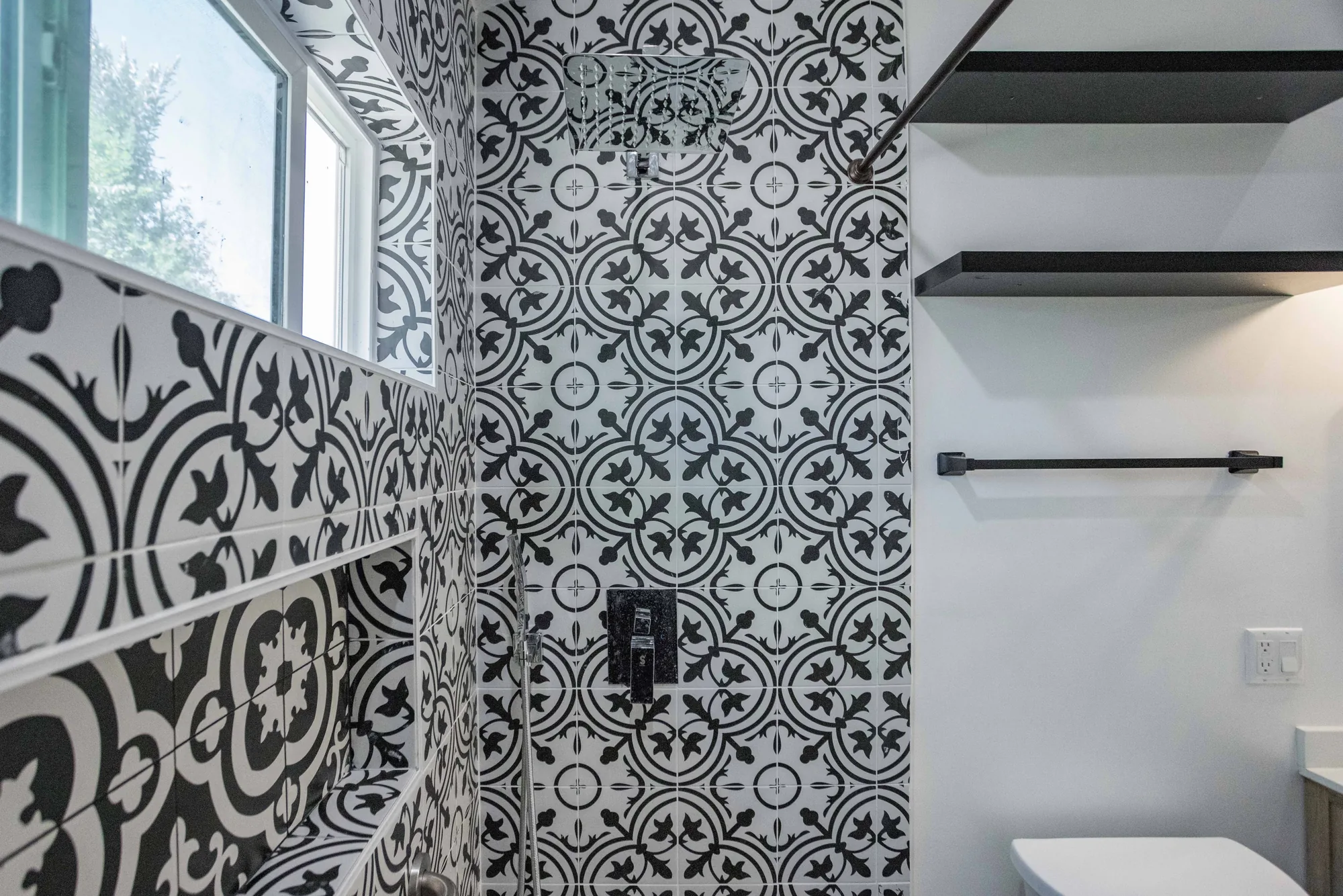Patterned tile shower wall with a black shower valve, black floating shelves, and a towel bar nearby.