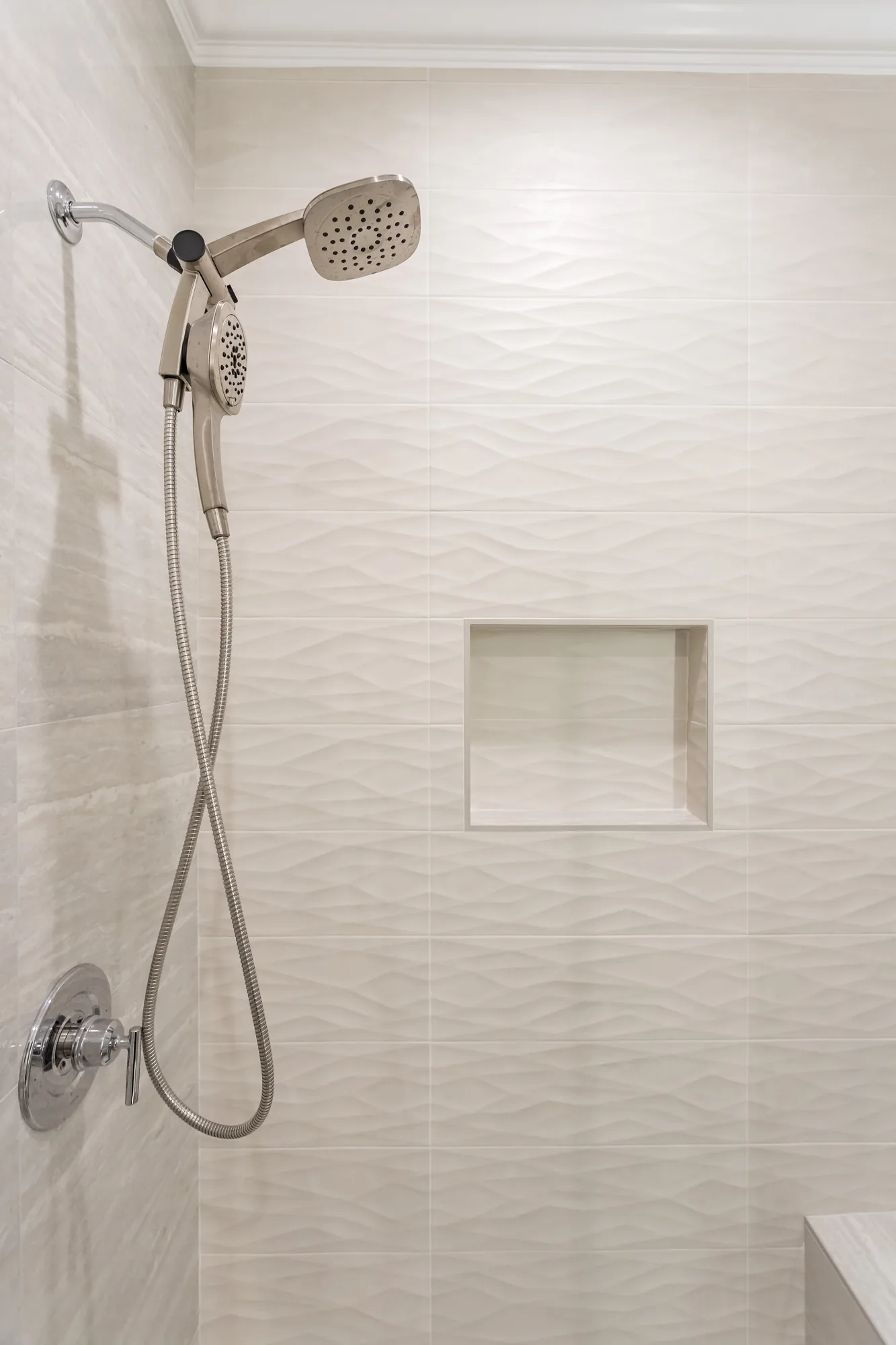 Chrome showerhead and handheld sprayer mounted on a wall of rippled white tile beside a recessed niche.