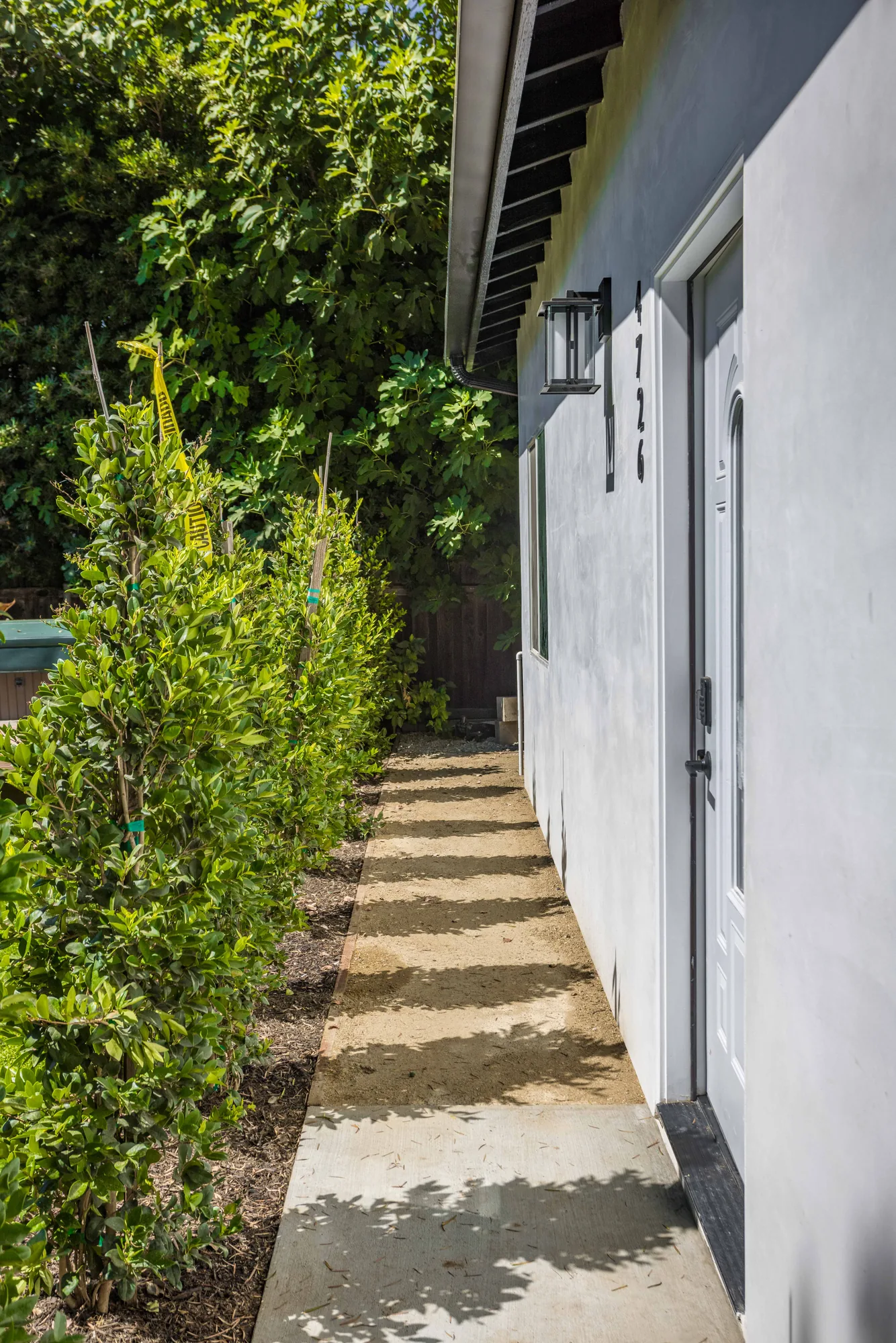 Narrow side yard walkway running past a hedge to the unit's white entry door and wall sconce.