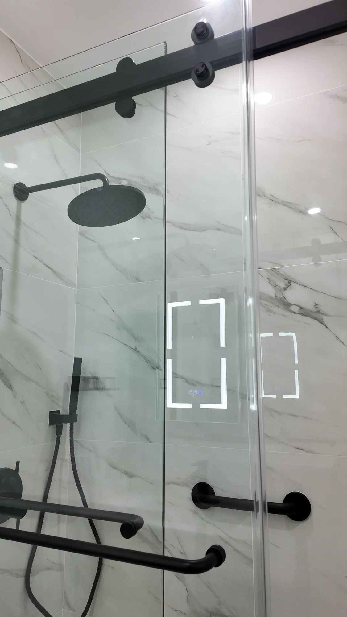 Sliding glass shower door on black roller hardware with a rain head and grab bars on marble-look tile.