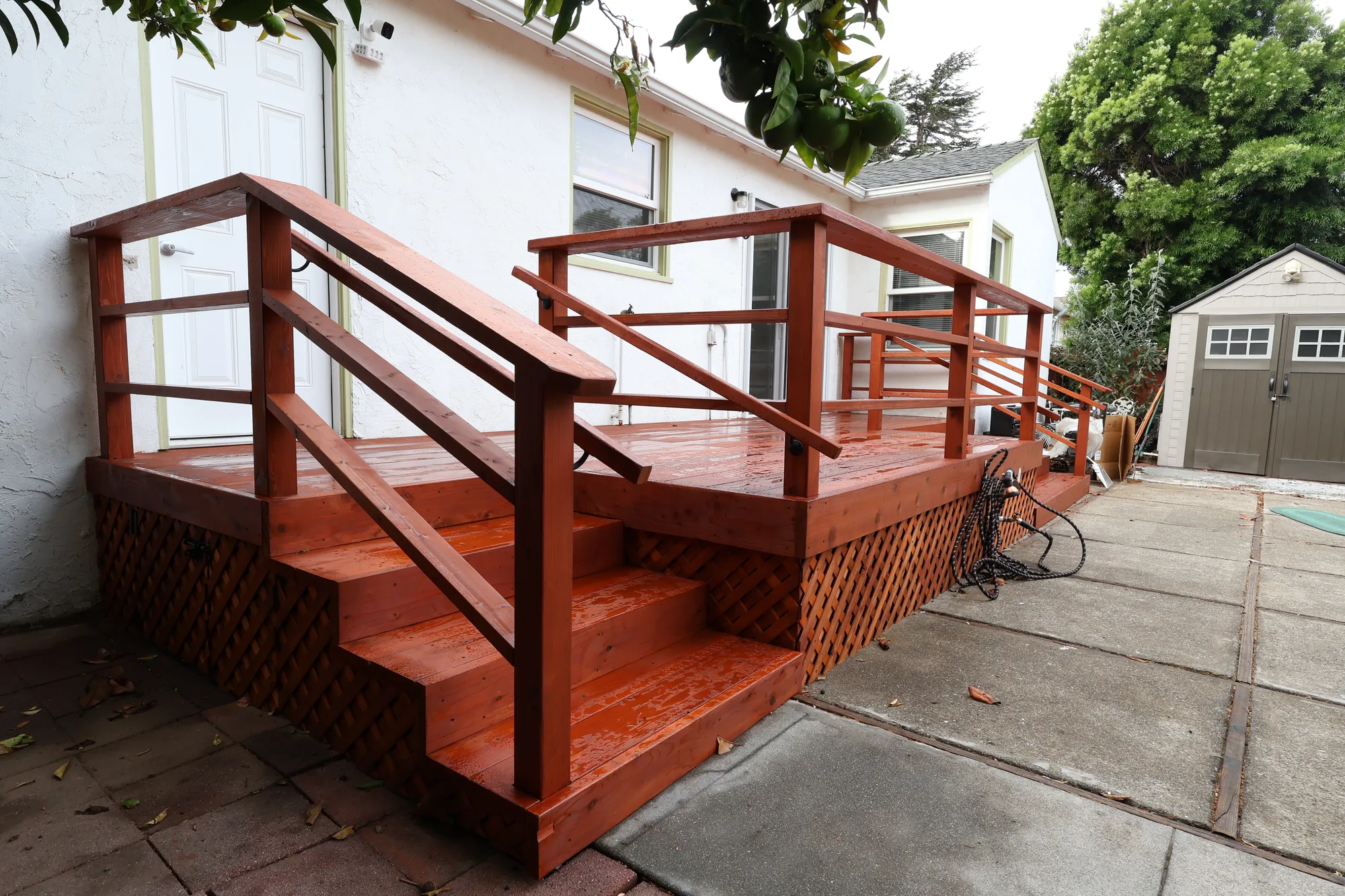 Corner view of a stained wood deck with open stair, wood handrail and lattice skirt beside a concrete patio.
