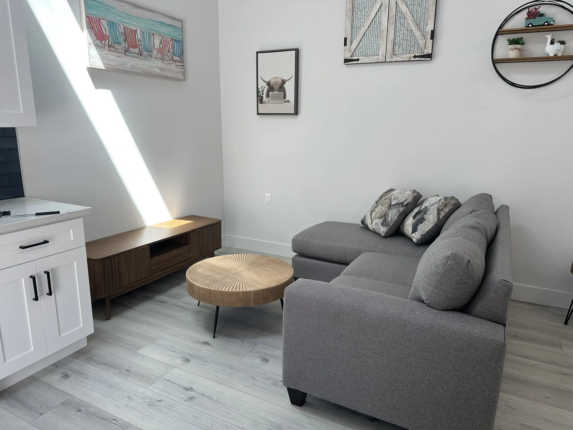 Living corner of a studio unit with gray sectional, round wood coffee table and walnut media console.