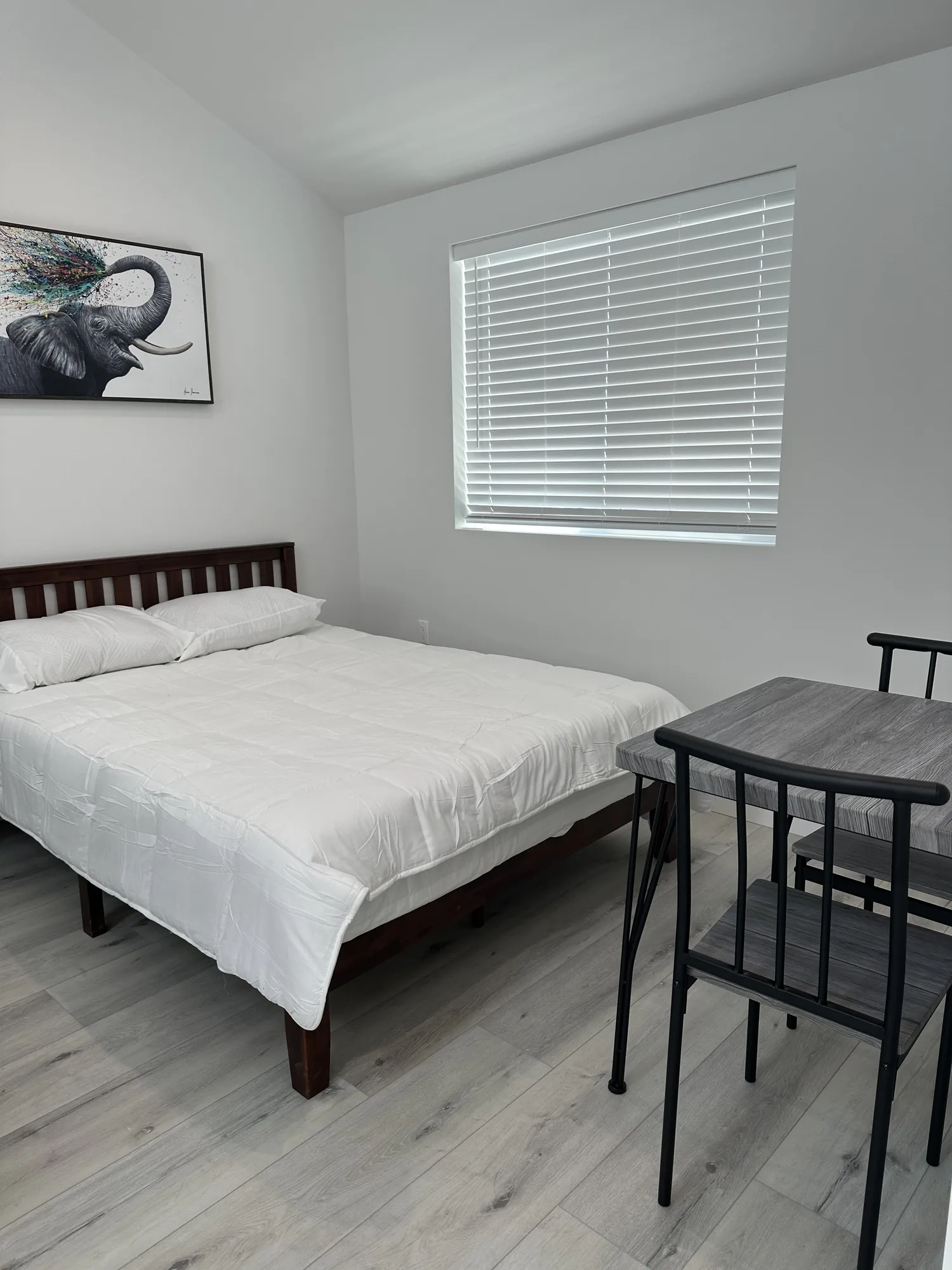 Sleeping area of a studio unit with a made bed, white window blinds and a small bistro table set.
