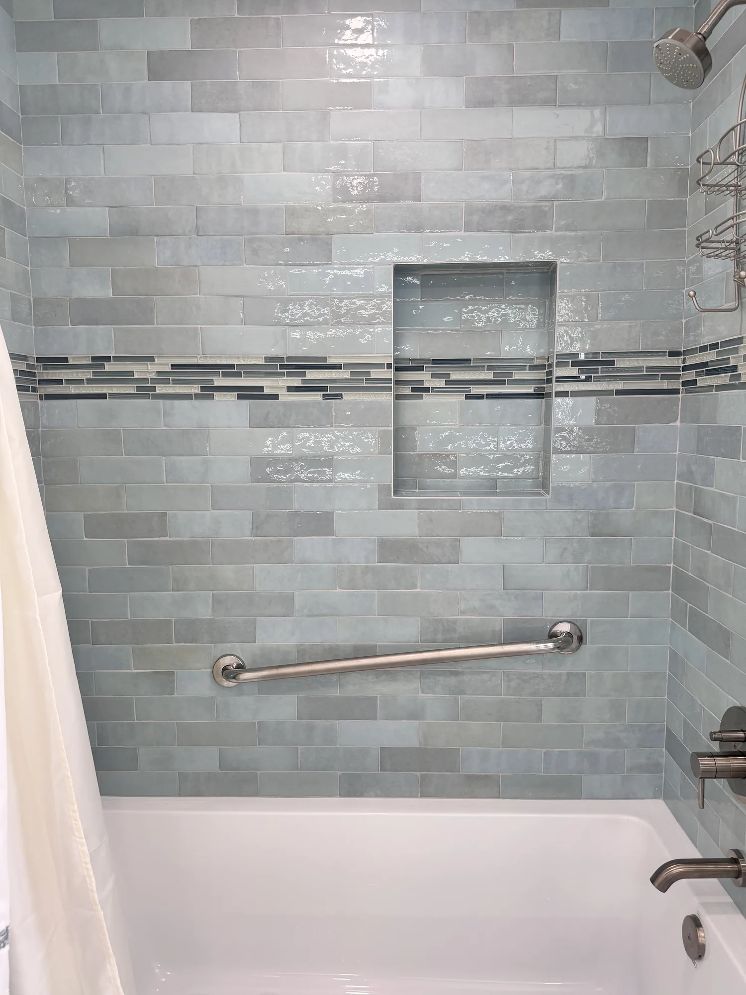 Tiled shower wall above the white tub with a recessed niche, mosaic stripe and grab bar.