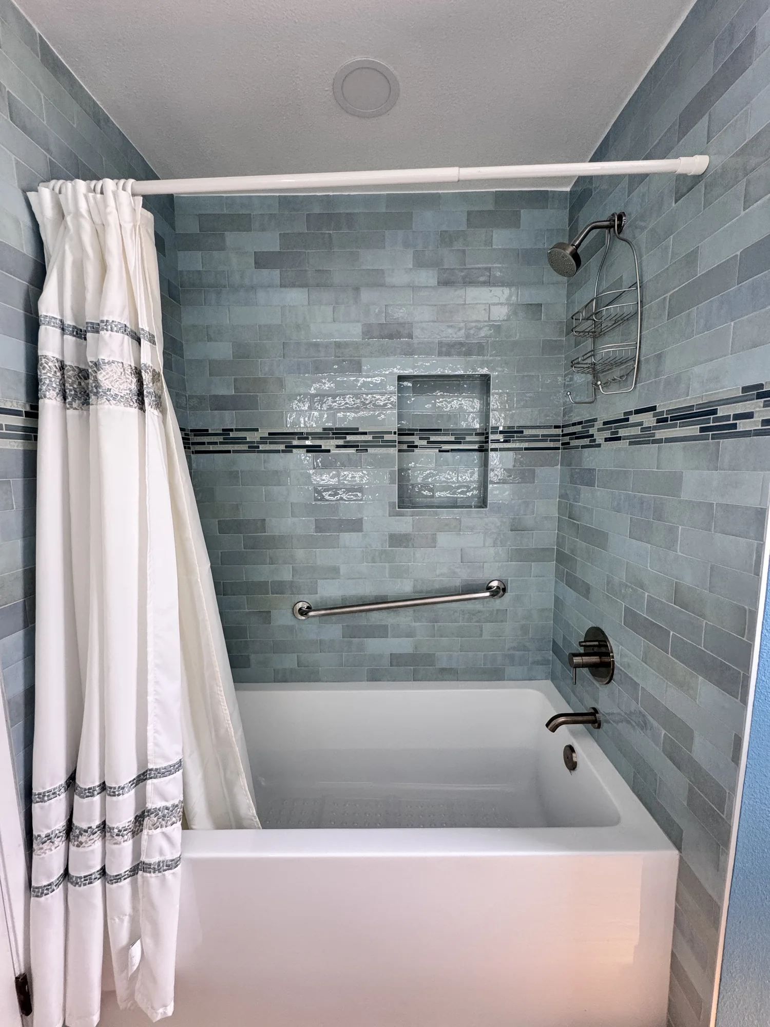 Remodeled tub and shower alcove with glossy blue-green tile, mosaic accent band and white curtain.