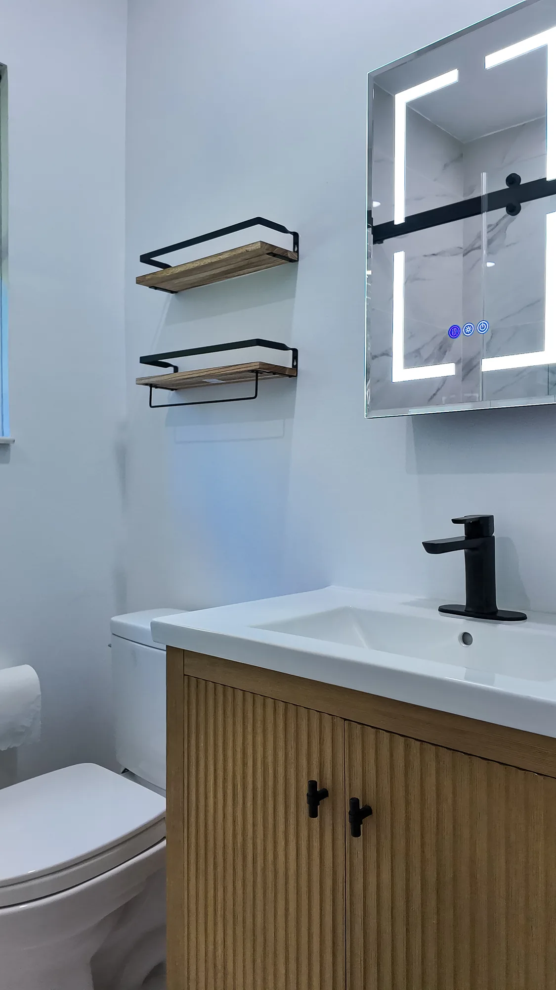 White toilet beside a vanity with black and wood wall shelves and a lighted mirrored cabinet above.