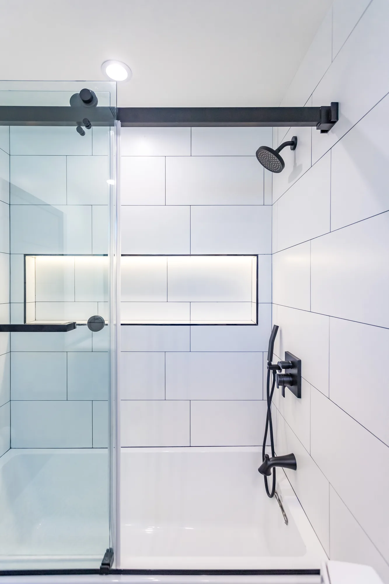 Tub shower with glass slider, matte black showerhead and handheld beside a backlit tile niche.