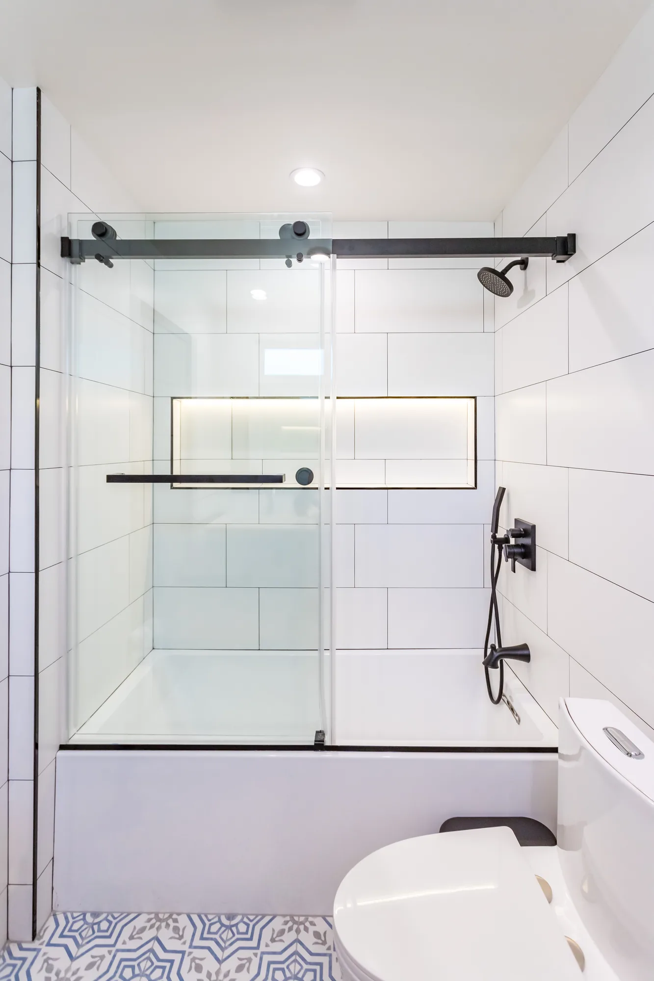 Tub shower with black-framed sliding glass door, lit tile niche and patterned blue floor tile.