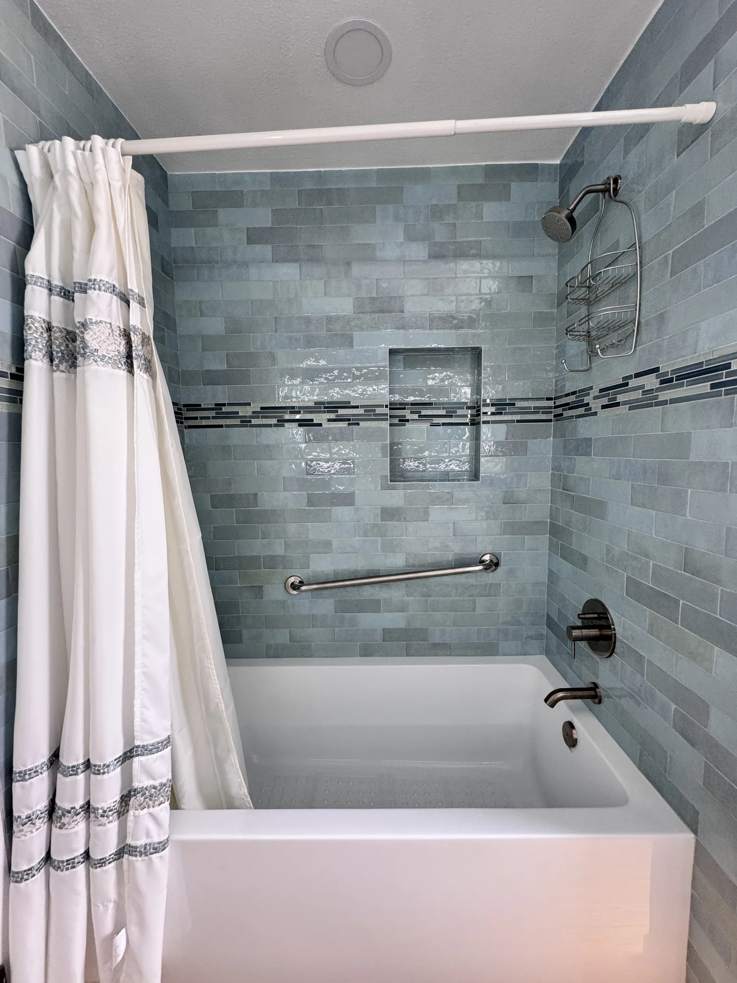 Tub and shower surround tiled in blue-green subway tile with a recessed niche and brushed nickel grab bar.