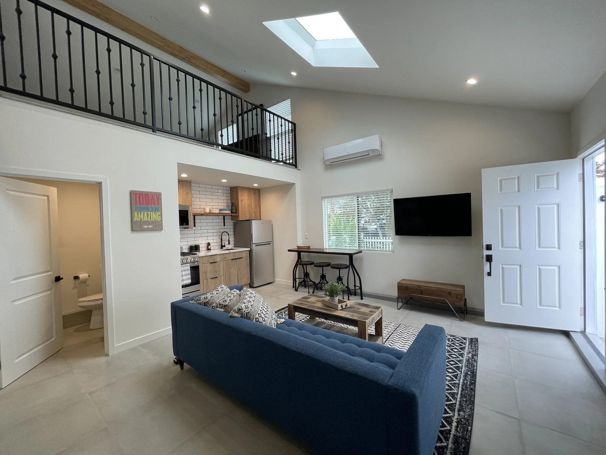 Wide great room of a two-story unit with skylight, loft railing, kitchen and open front door.