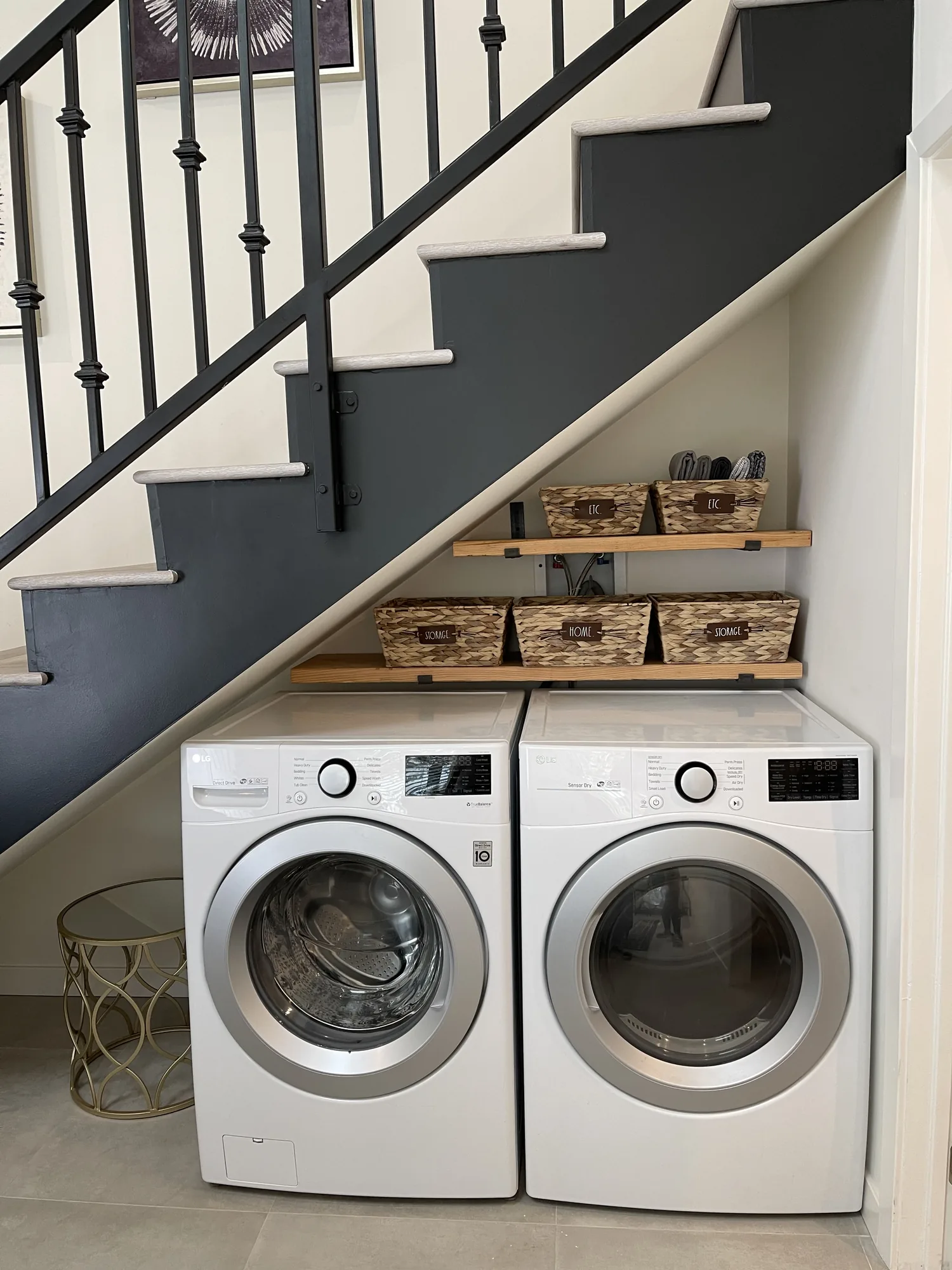 Front-load washer and dryer tucked under a staircase with two wood shelves and baskets above.