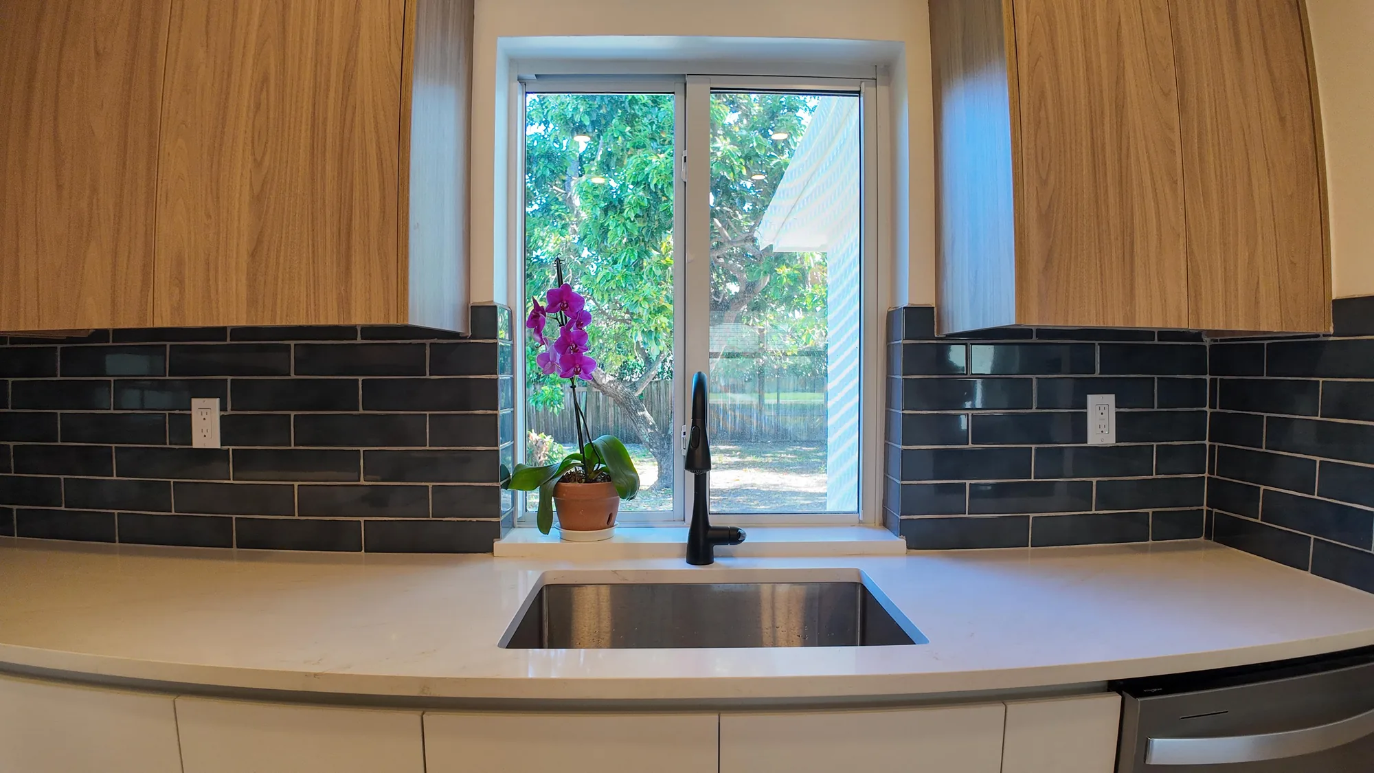 Undermount sink below a window with a matte black faucet, light quartz counter and dark tile on both sides.