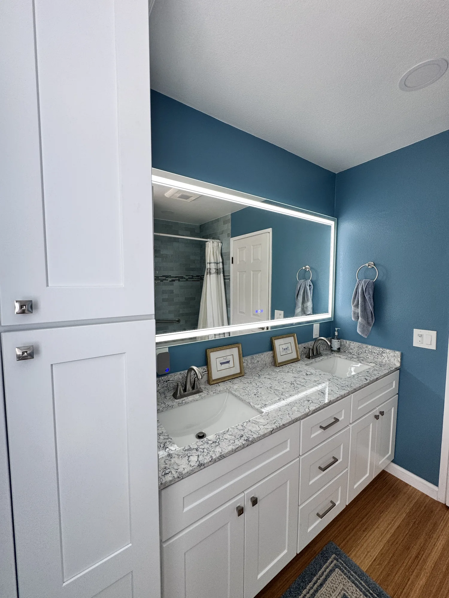 Angled view of the double vanity, backlit mirror and tall white linen cabinet in a blue bathroom.