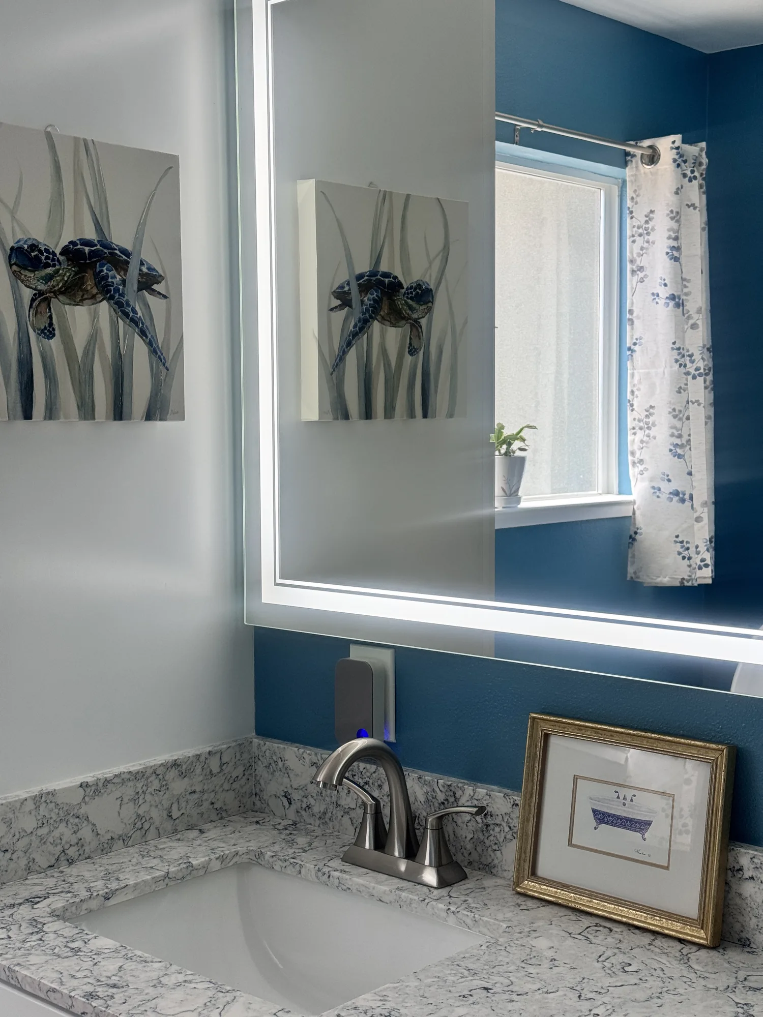 Corner of the vanity where the lit mirror reflects framed artwork and the curtained privacy window.