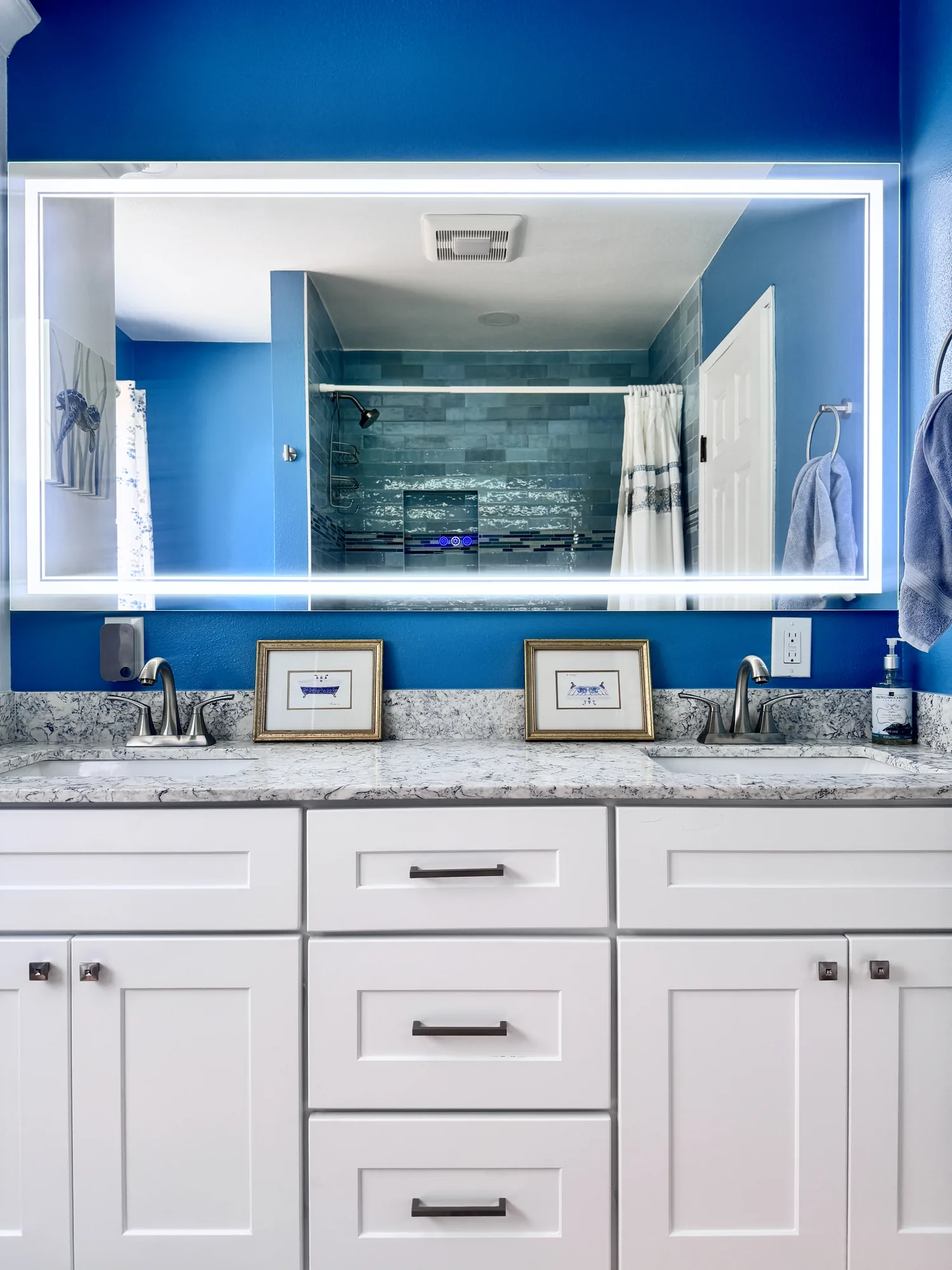 Backlit mirror above a white double vanity reflects the tiled shower across the bathroom.