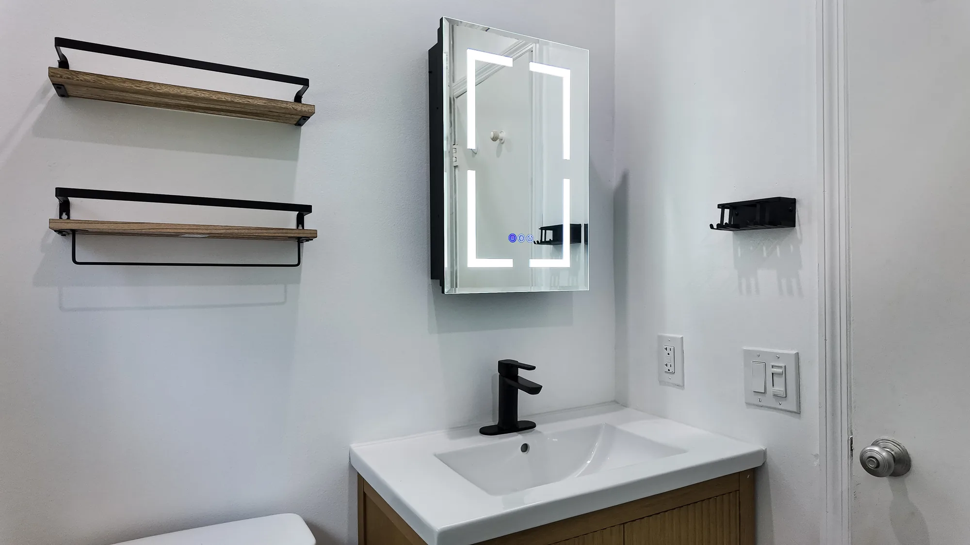 Vanity wall with wood and black open shelves, a lighted mirrored cabinet and a white integrated sink.