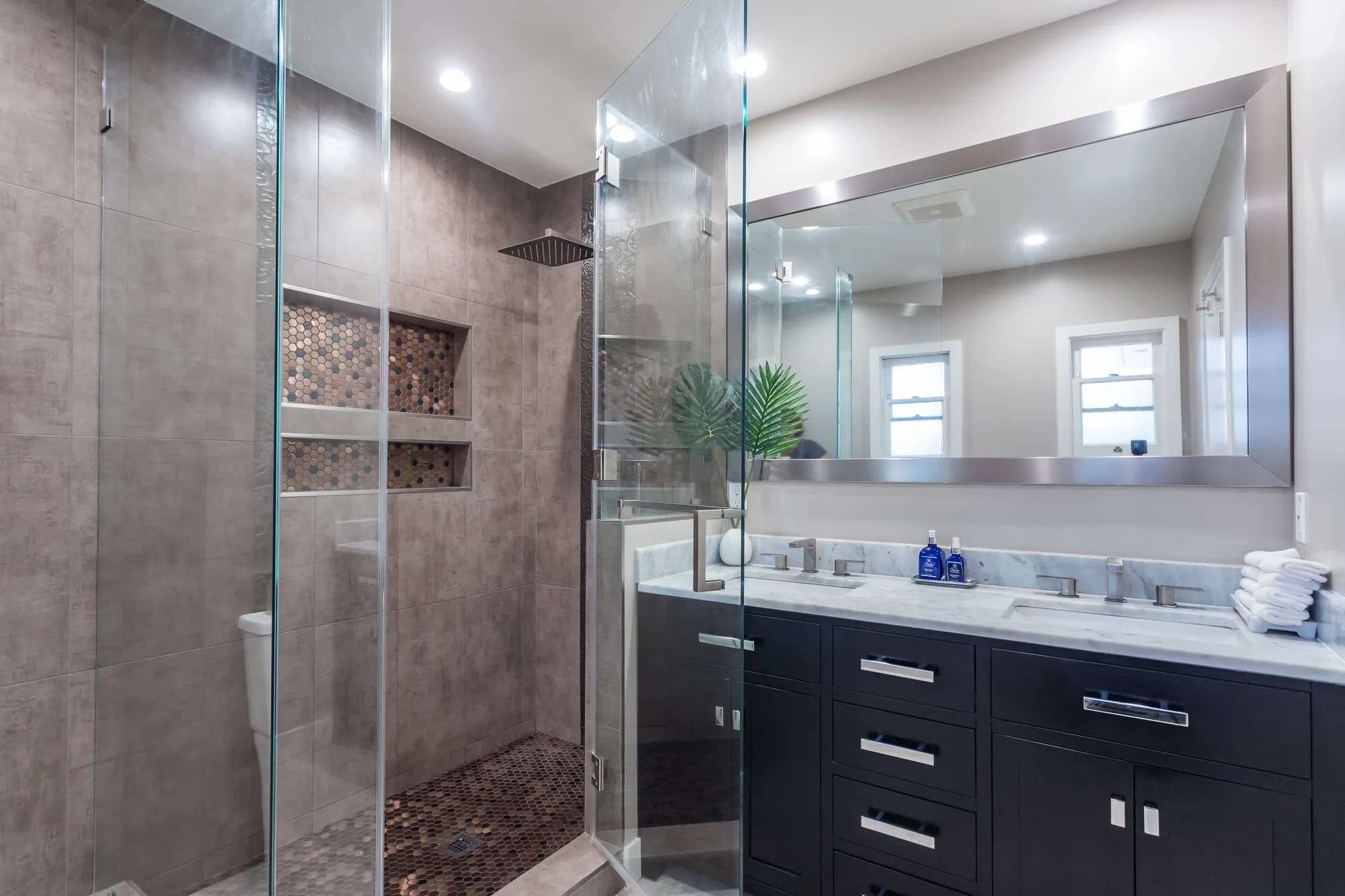 Wide view of a remodeled bathroom with a walk-in glass shower and dark double vanity under a mirror.