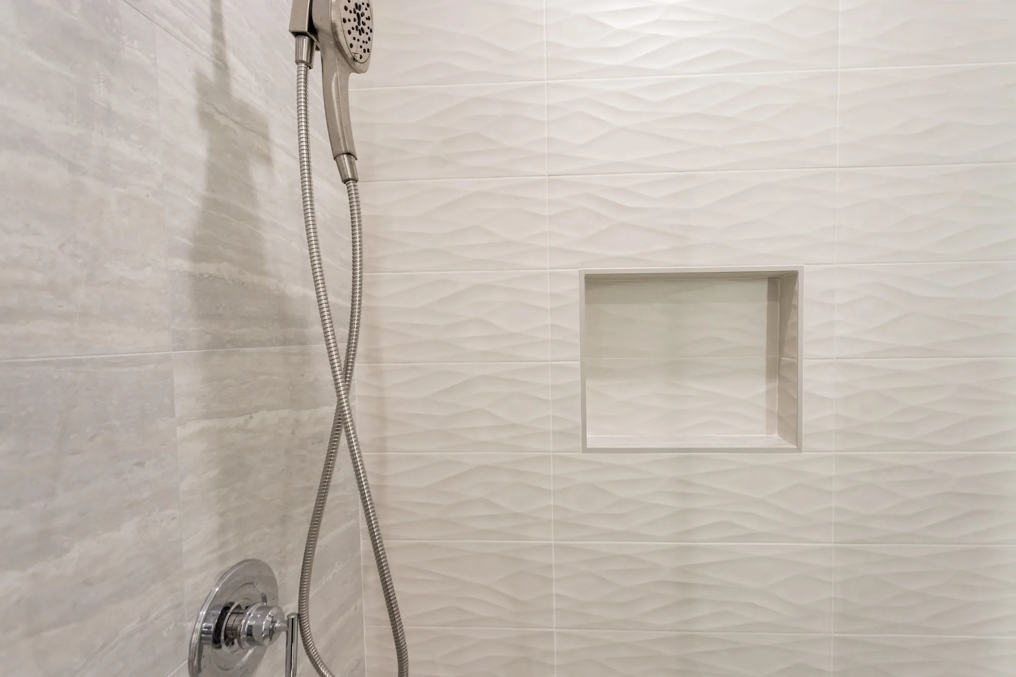 Close-up of wavy textured white shower tile with a recessed niche and a chrome handheld shower fixture.