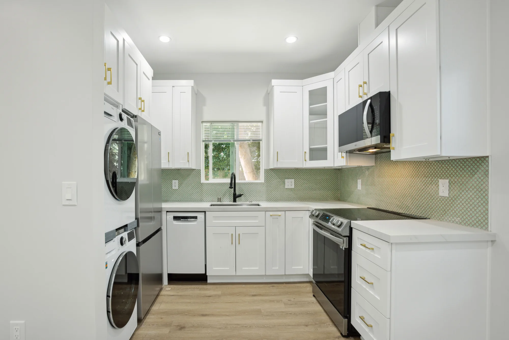 Small white shaker kitchen with green tile backsplash, brass pulls and a black faucet at the window.