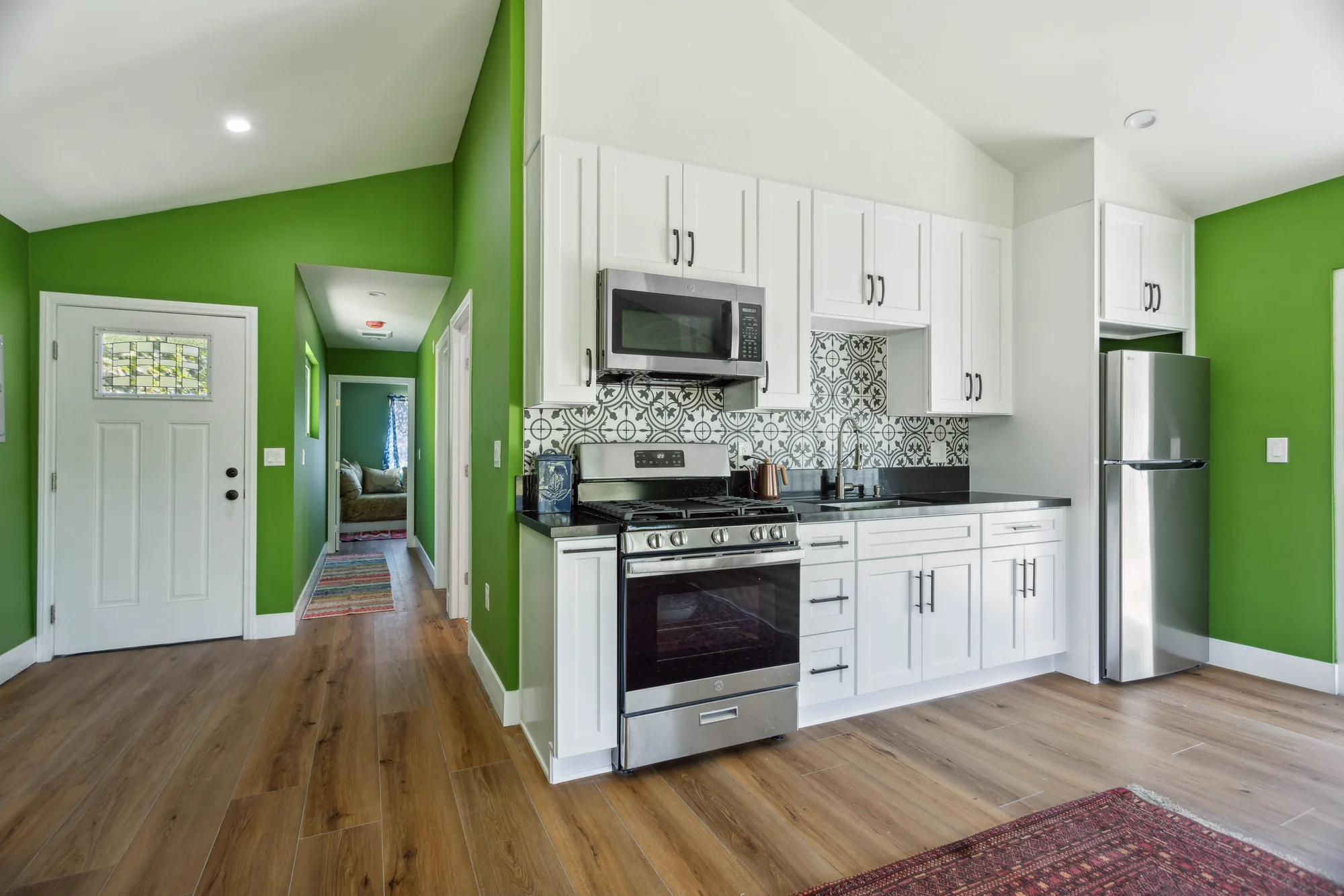 White shaker kitchen cabinets with dark counters and a bold patterned tile backsplash beside a hallway.