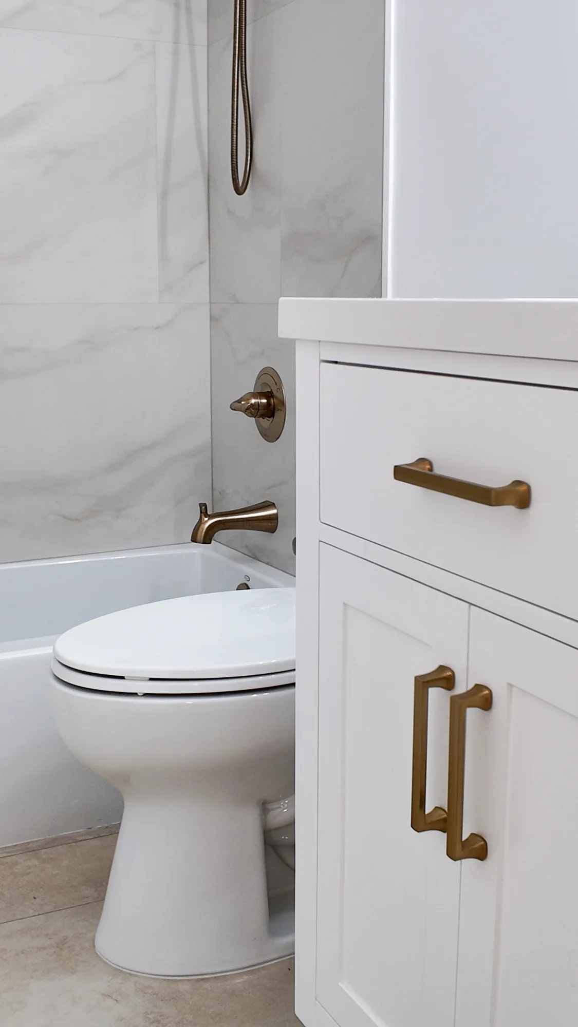 White shaker vanity with gold pulls next to a toilet and a marble-look tiled tub and shower alcove.