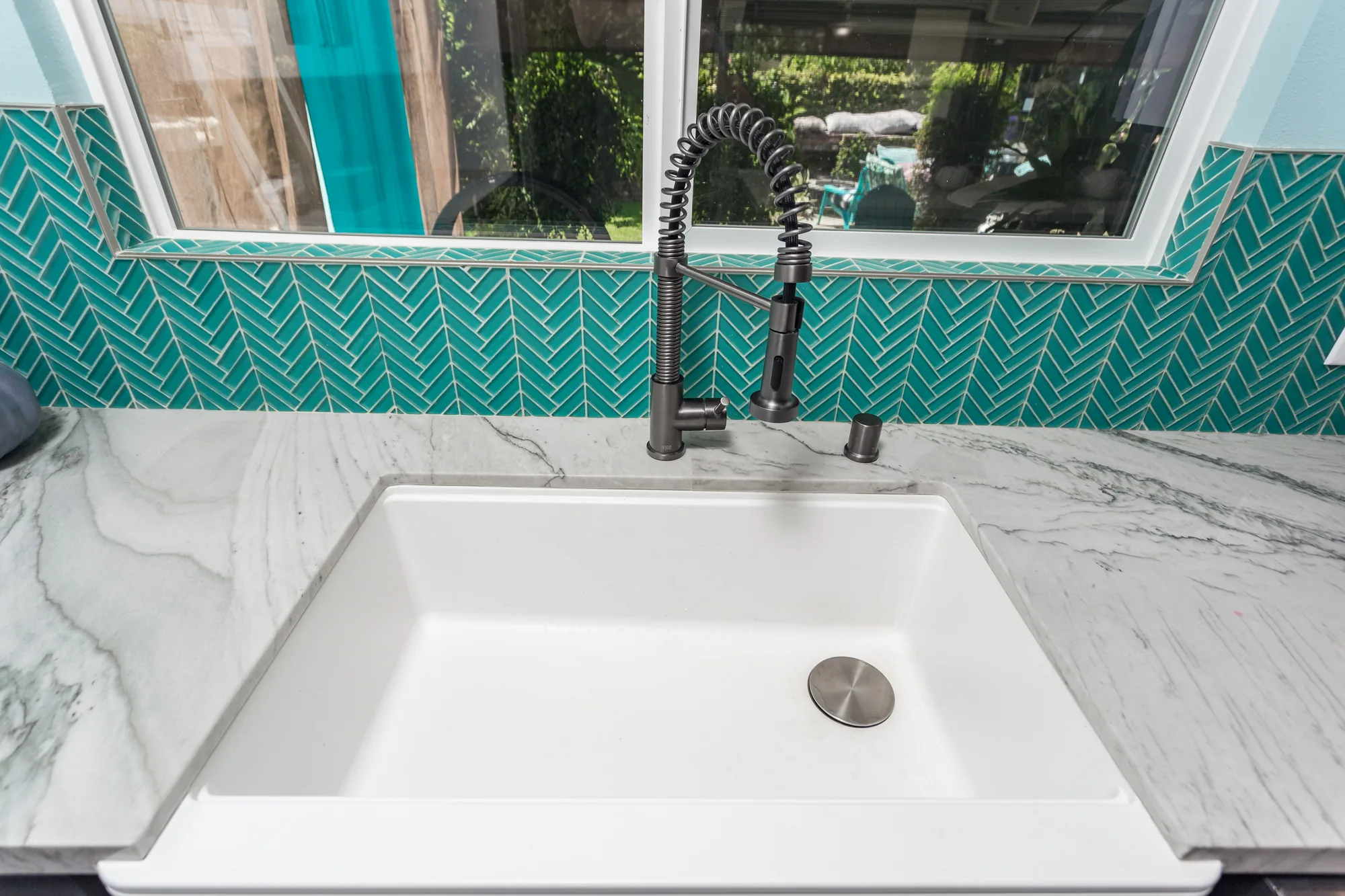 Detail of a white single-bowl sink with a dark coil-spring faucet against teal herringbone tile.
