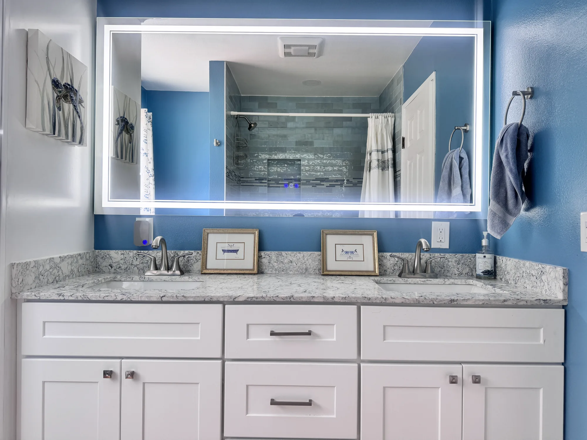 Wide view of a white double vanity with stone-look counter, backlit mirror and deep blue walls.